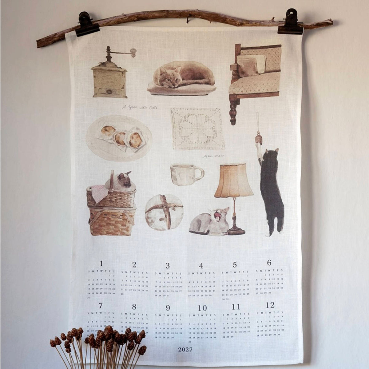 A fabric wall calendar hanging from a rustic wooden rod with black clips, featuring vintage-style illustrations of cats, a coffee grinder, a lamp, and a calendar grid for 2027. The calendar is displayed against a plain off-white wall with dried flowers at the bottom.