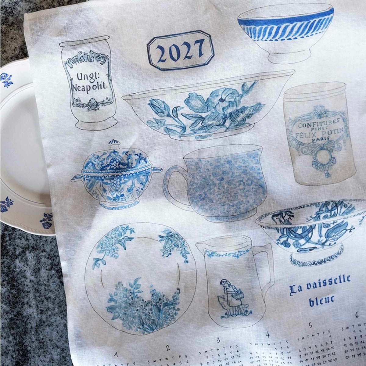 A white linen fabric tea towel printed with blue vintage ceramic motifs, including a teapot, cups, plates, and a calendar at the bottom. It is laid flat on a dark stone surface with partial white dishes visible at the edges.