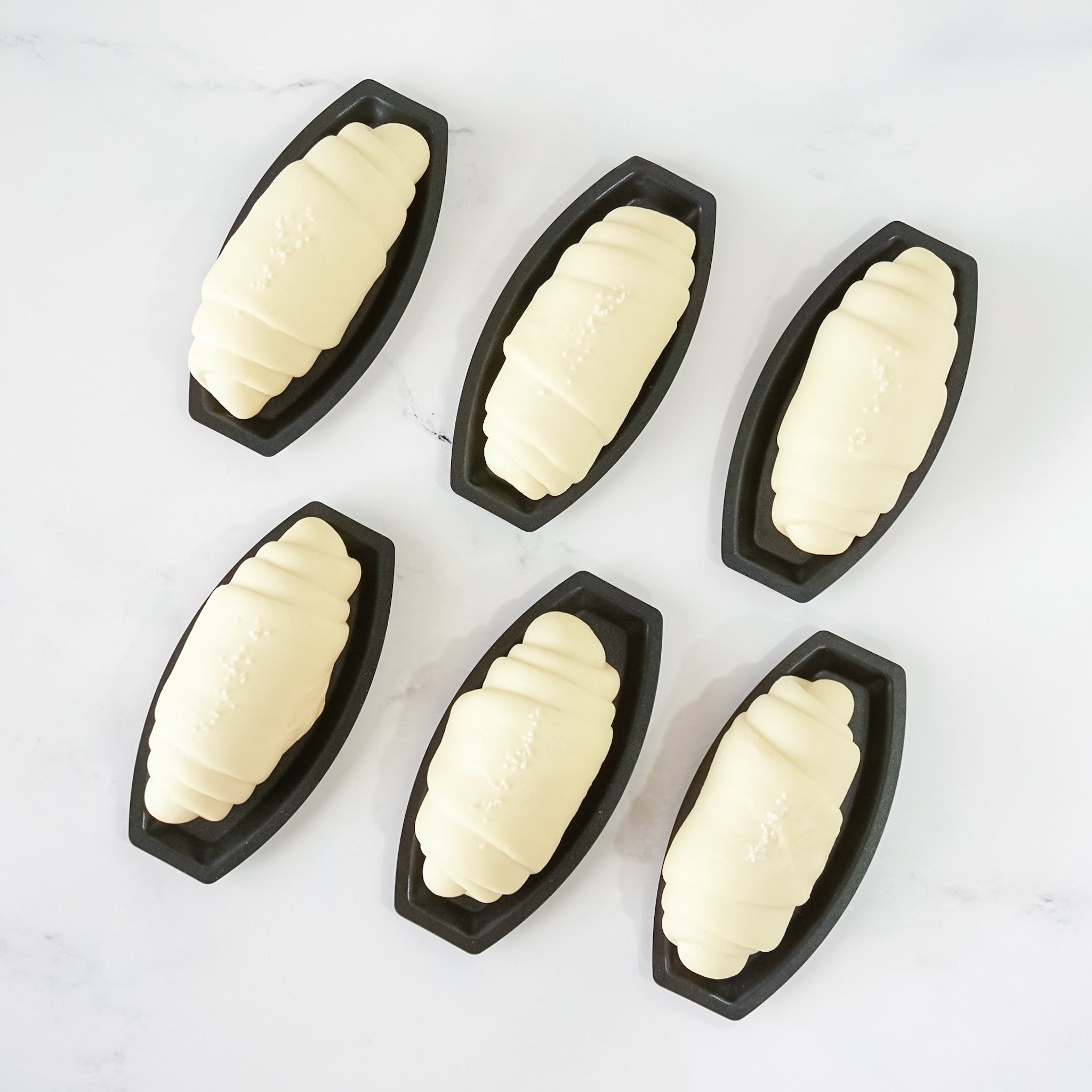 Six black boat-shaped molds with butter roll dough inside on a white background