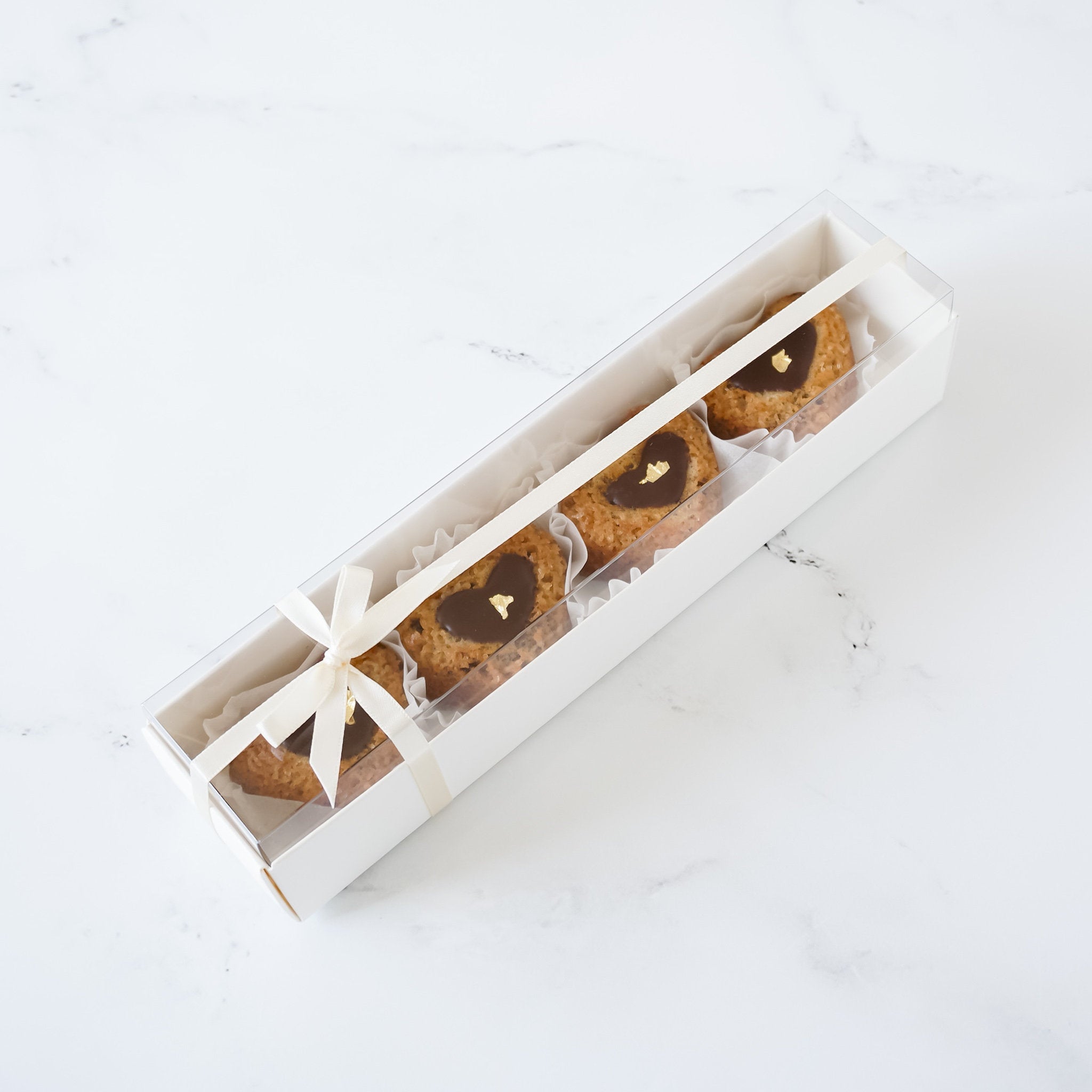 A white box with a clear lid containing four heart-shaped cookies. The cookies are topped with chocolate and a gold leaf.