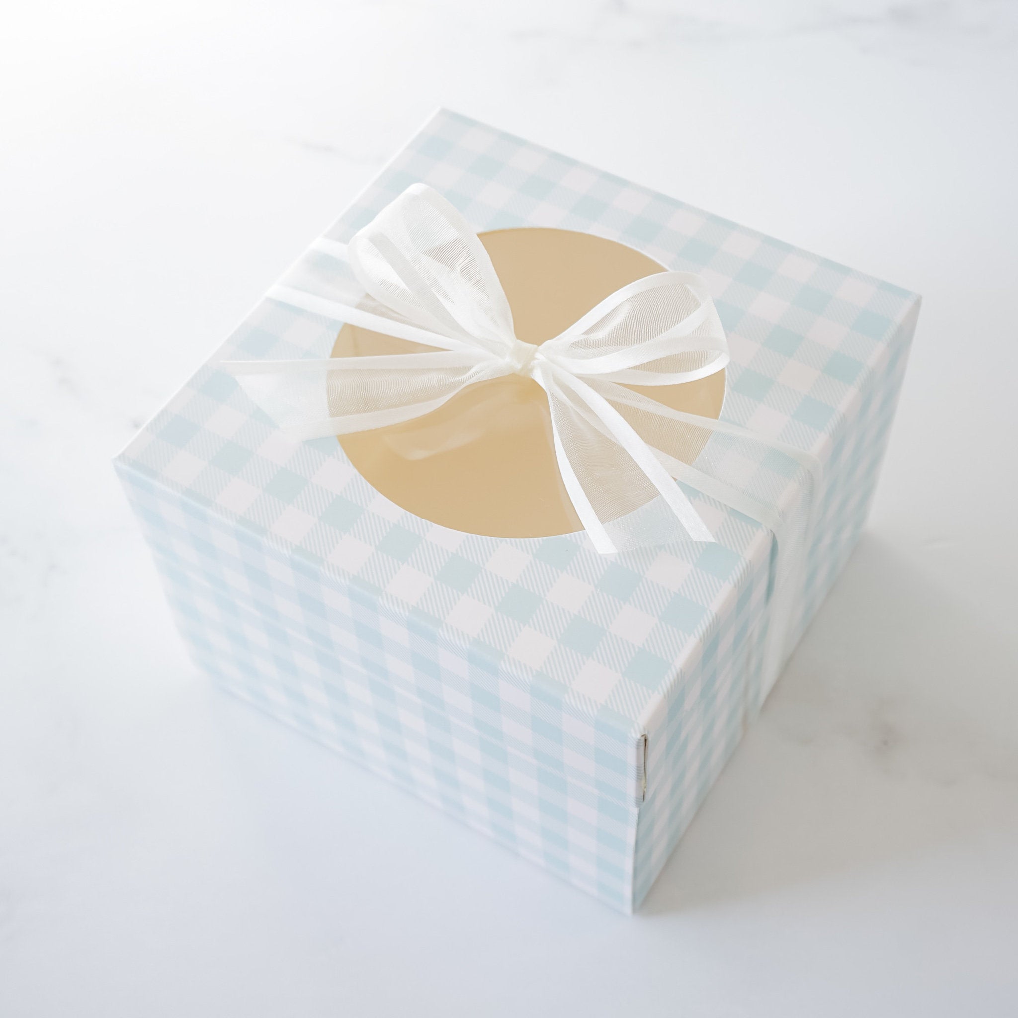 A light blue and white gingham gift box with a white sheer ribbon tied in a bow. The box has a circular window in the top. The box is on a white marble surface.