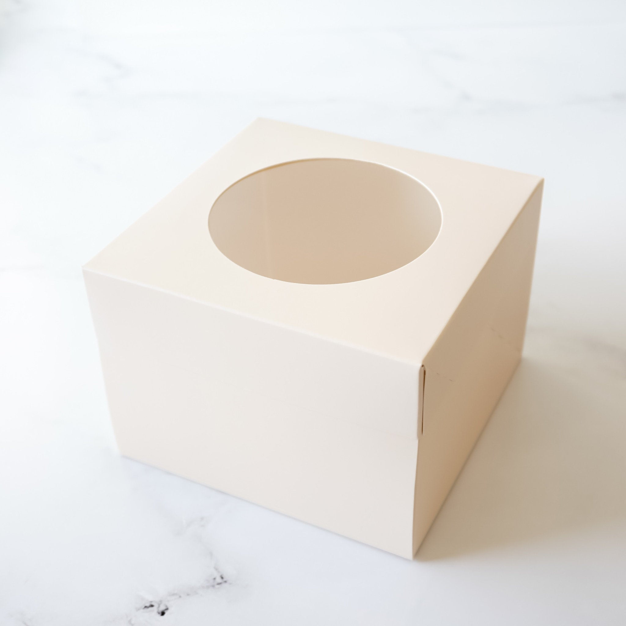 An ivory cardboard box with a circular window cut out of the top. The box is designed for packaging cupcakes or other small treats.