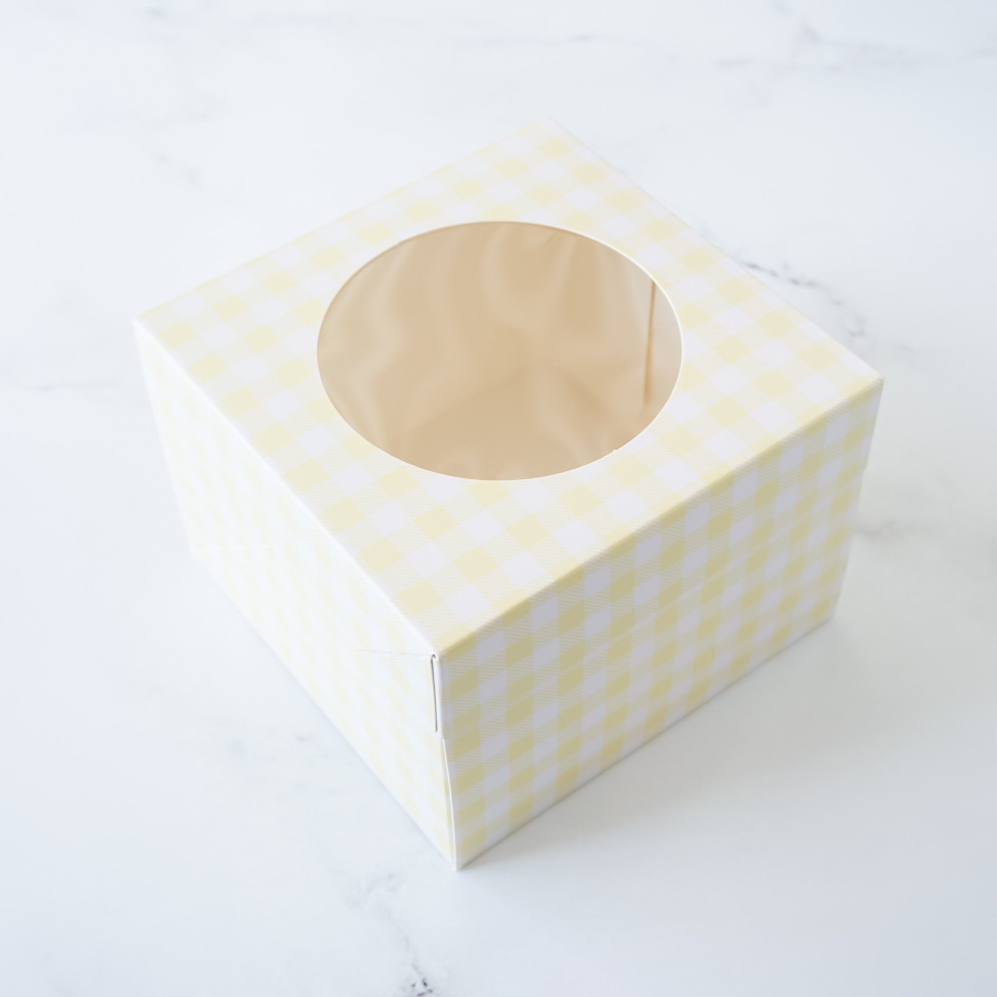 A yellow and white checkered cardboard box with a clear window on the top. The box is designed to hold a single cupcake or other small treat.