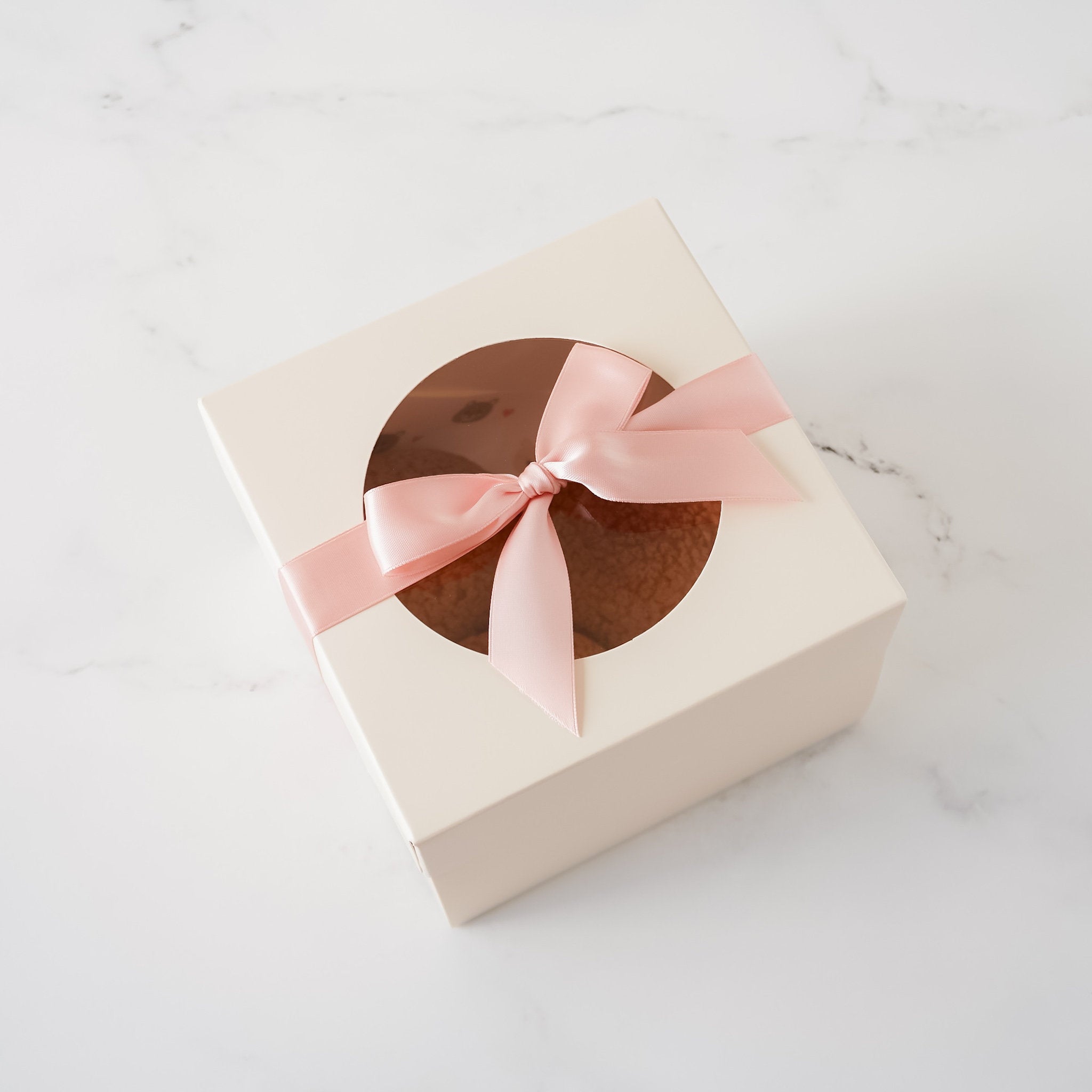 An ivory cookie box with a round window on top, adorned with a pink ribbon, against a marble background.