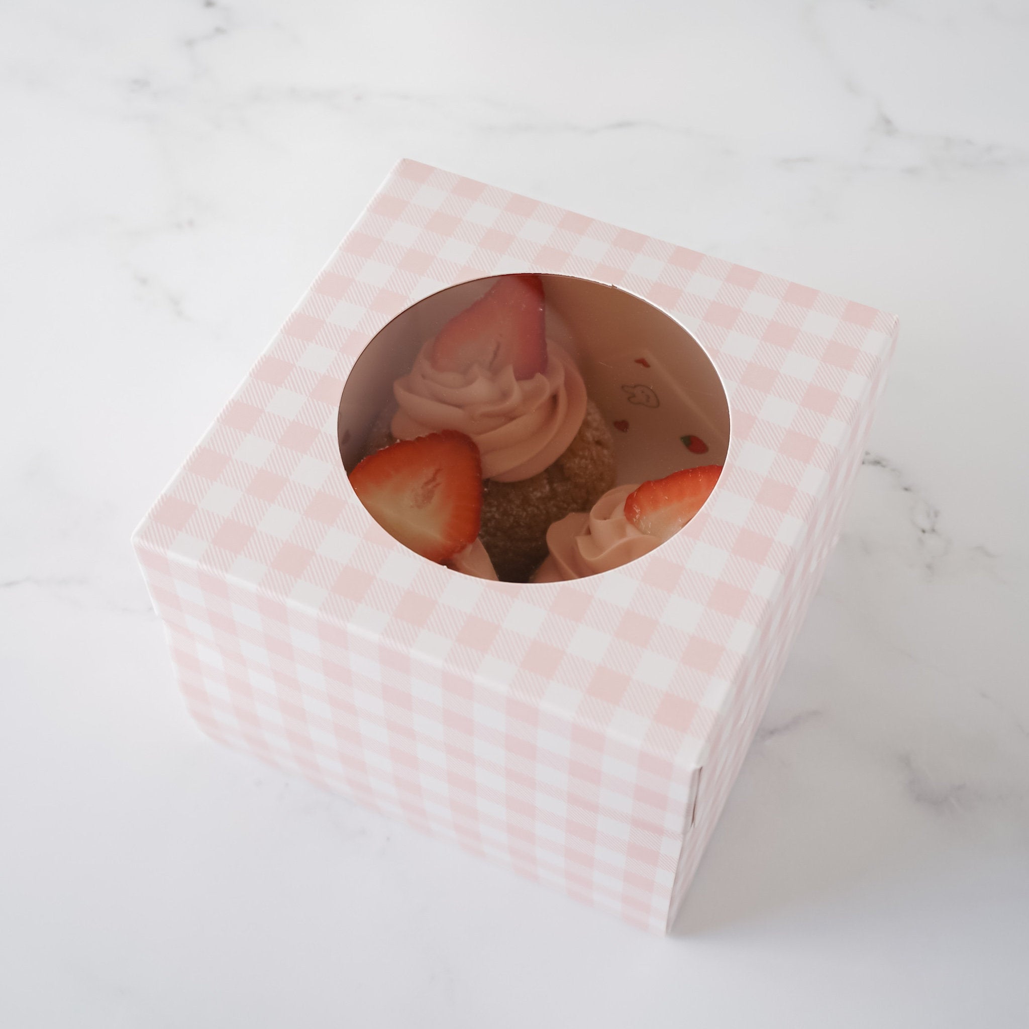 A pink and white gingham gift box with a clear window. The window shows three cupcakes with pink frosting and strawberries on top.