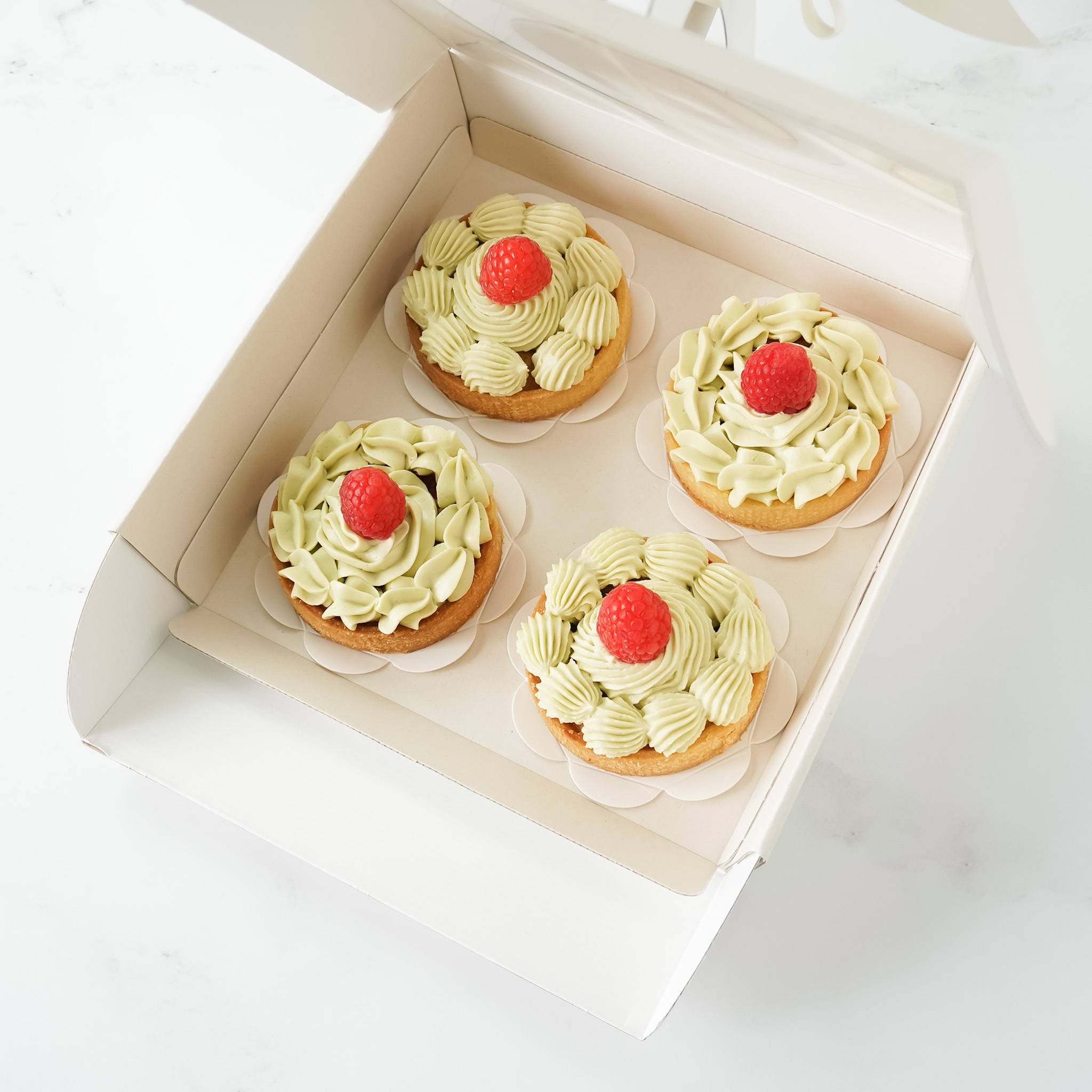Four small green and white tarts with a single red raspberry on top, arranged in a light beige.