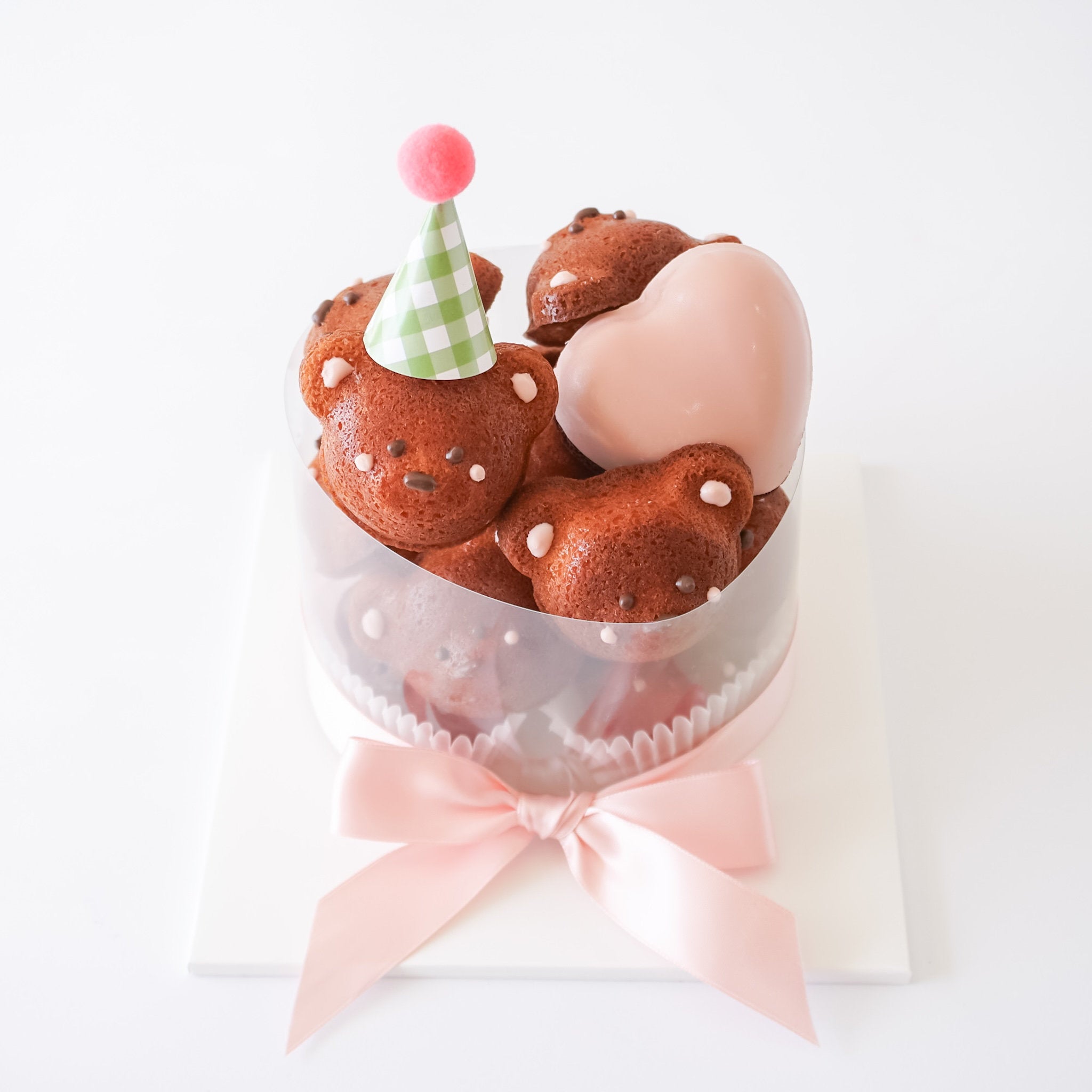 A round clear plastic cupcake container filled with vanilla madeleines shaped like teddy bears, topped with a pink heart-shaped decoration and a green-and-white checkered party hat. The container is wrapped with a pale pink ribbon and tied in a bow on a white square base against a white background.