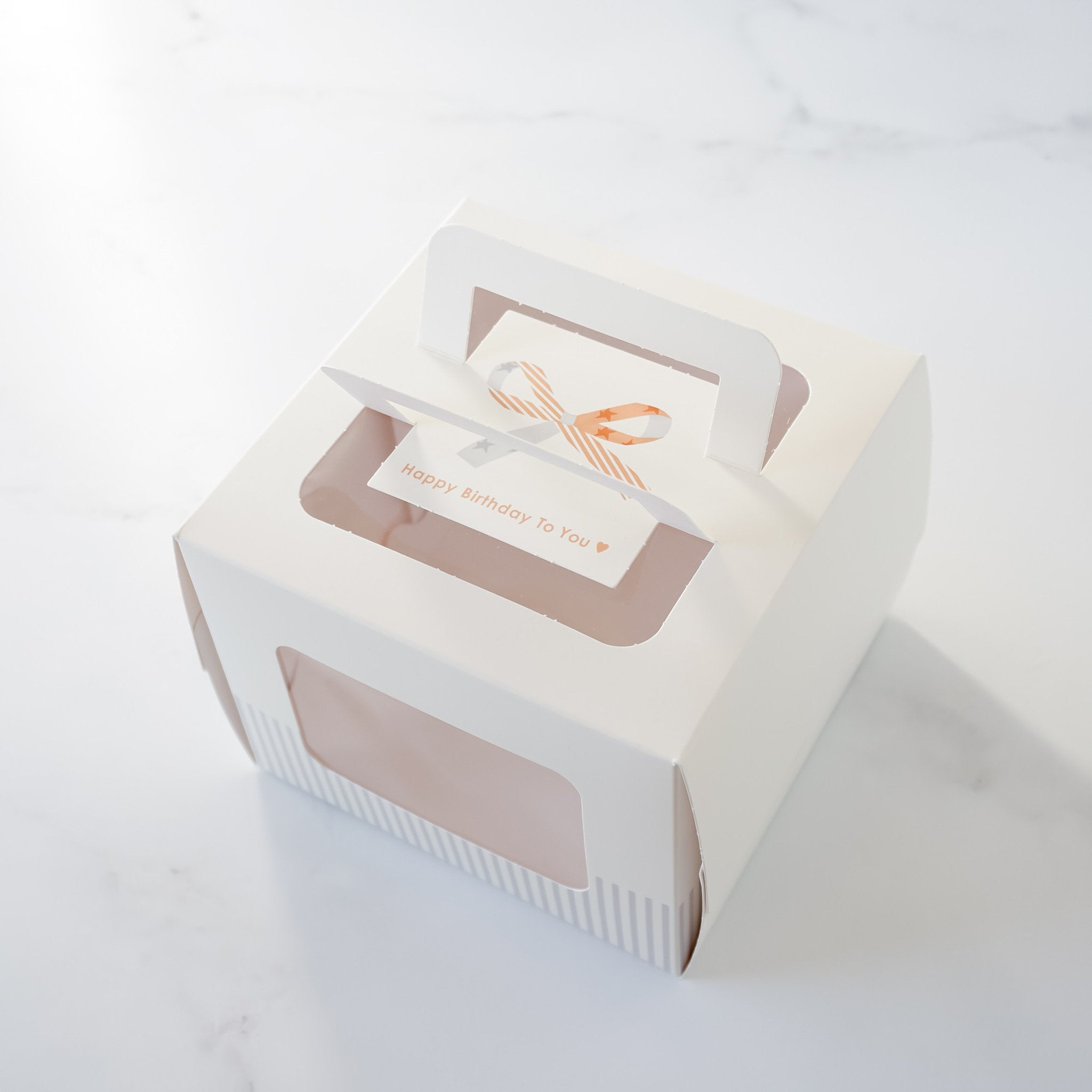 A white cardboard cake box with a window on the top. The window has a decorative brown and white ribbon design. The box has a striped pattern on the bottom and two handles on the sides.