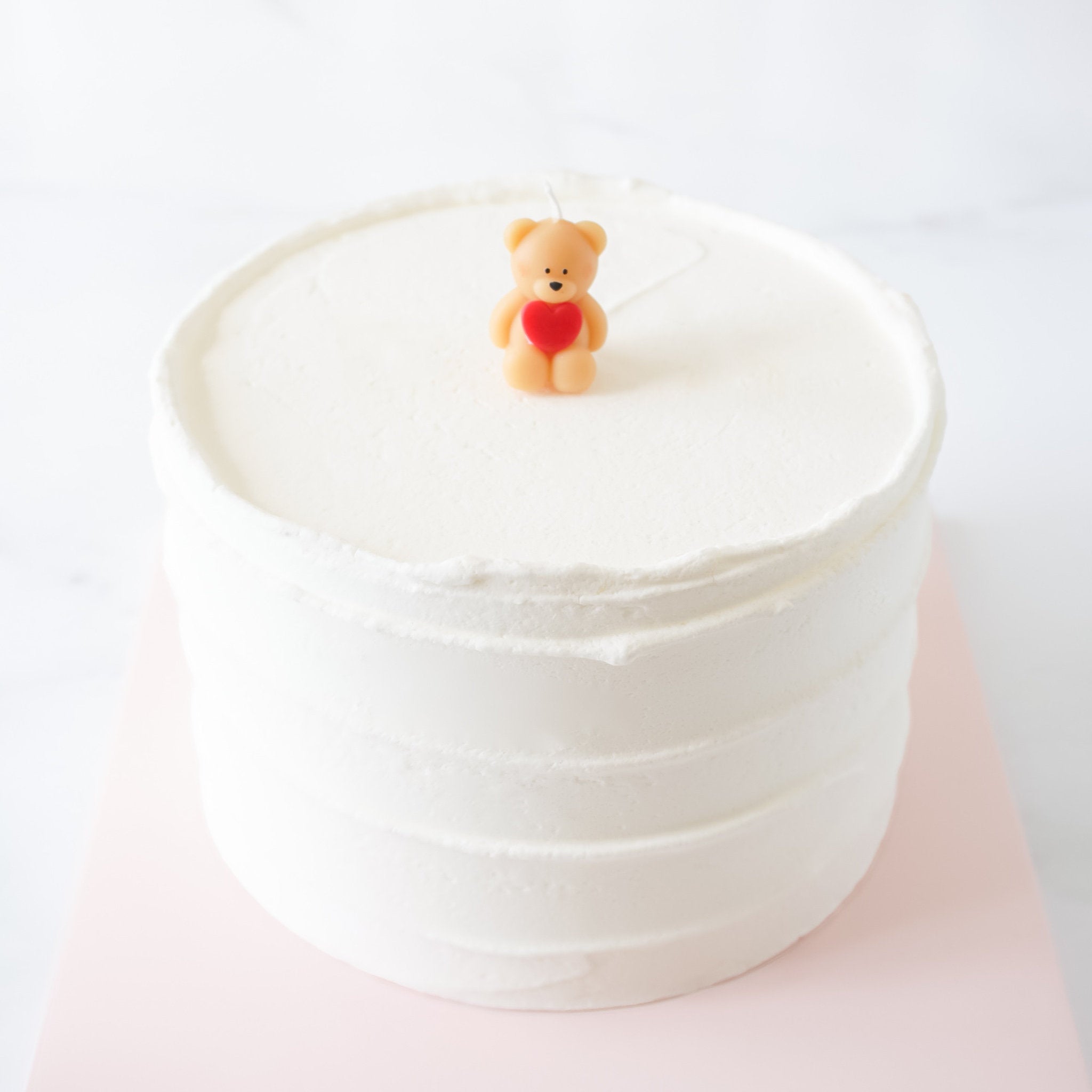 A white birthday cake topped with a miniature bear candle holding a red heart on a pink and white background.