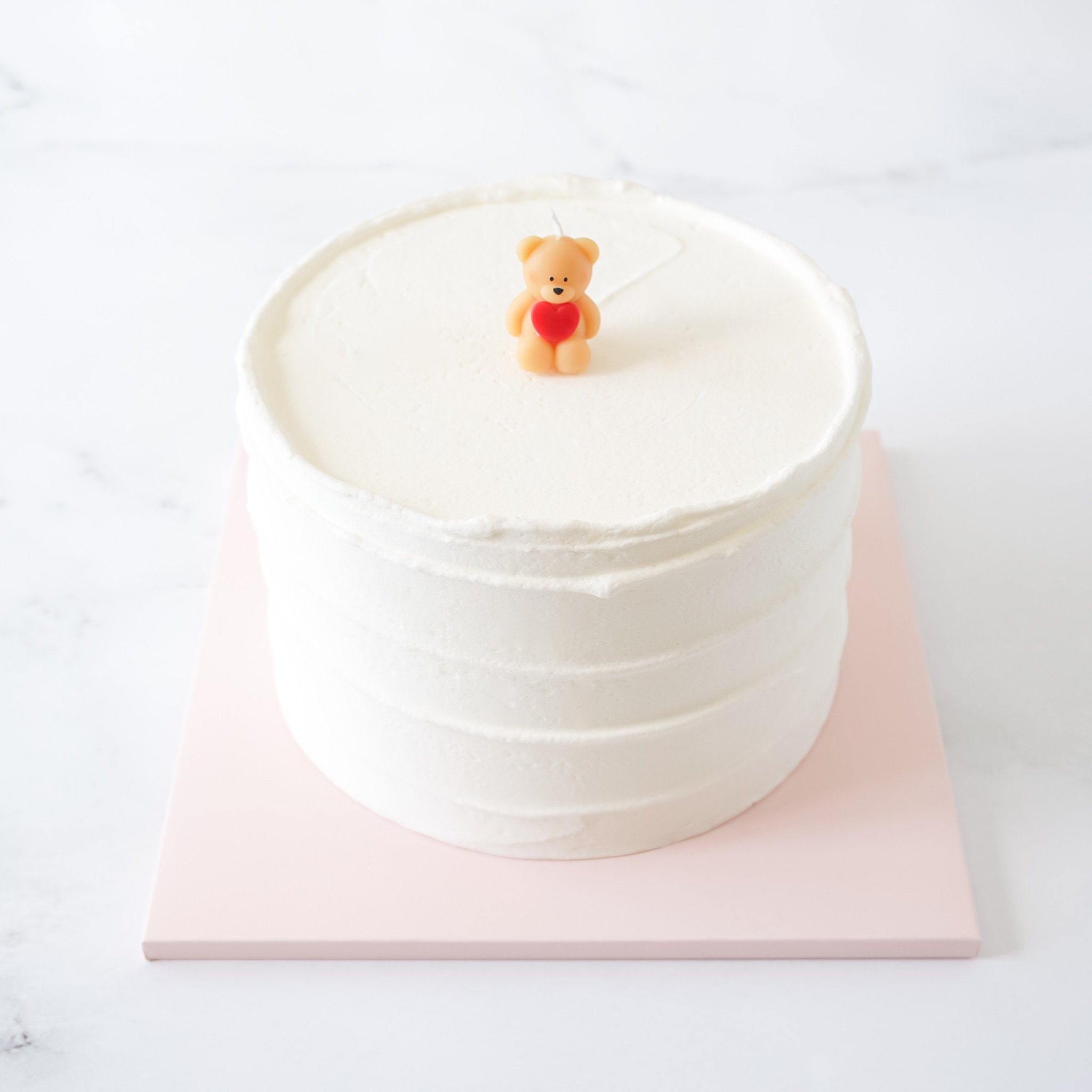 A white birthday cake topped with a miniature beige bear candle holding a red heart on a pink and white background.
