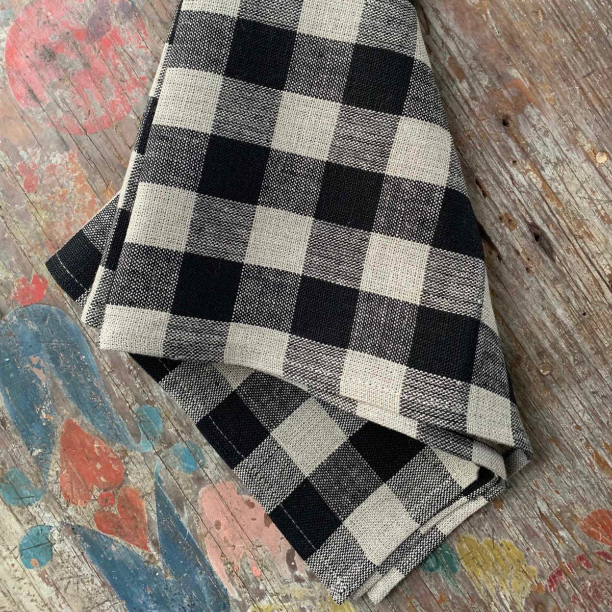 A folded black and off-white buffalo plaid fabric item, likely a napkin or small towel, displayed on a rustic, weathered wooden surface with colorful paint splatters.