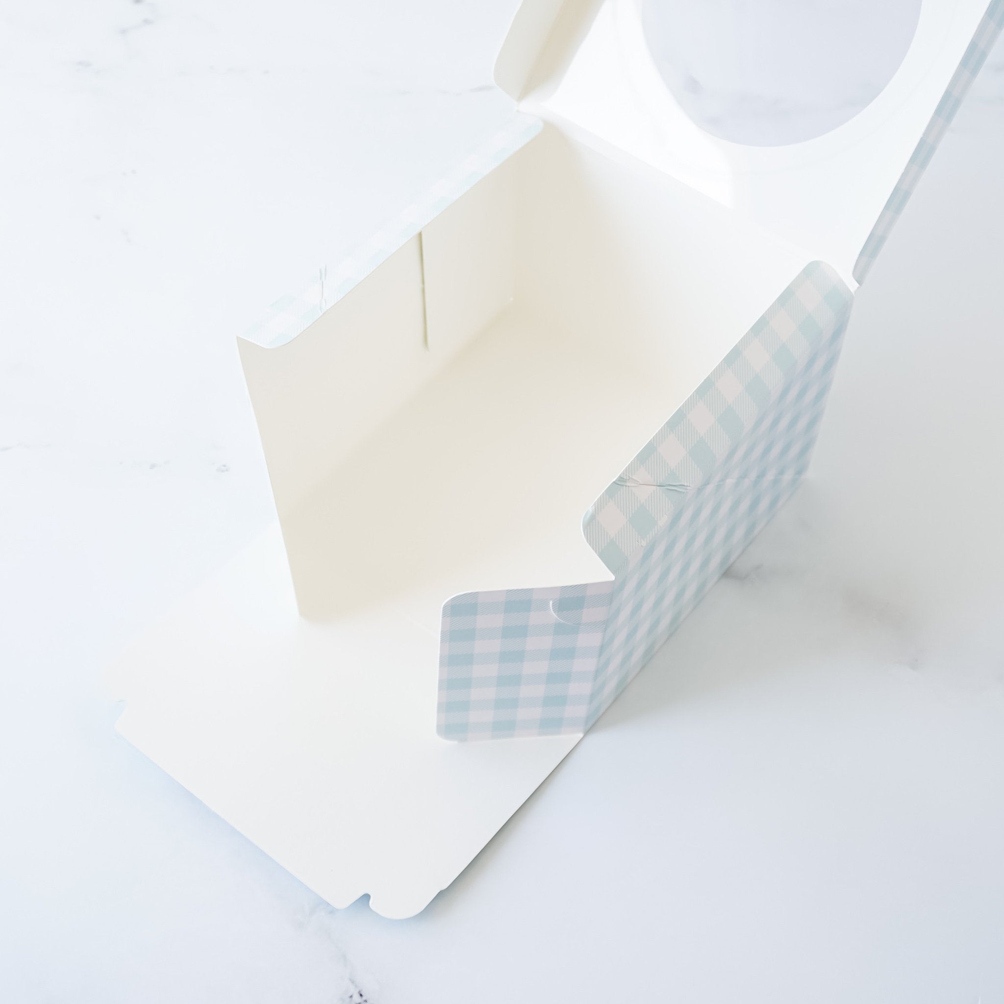 A white cardboard box with a blue and white gingham pattern. The box has a clear window on the top and is open to reveal the inside.