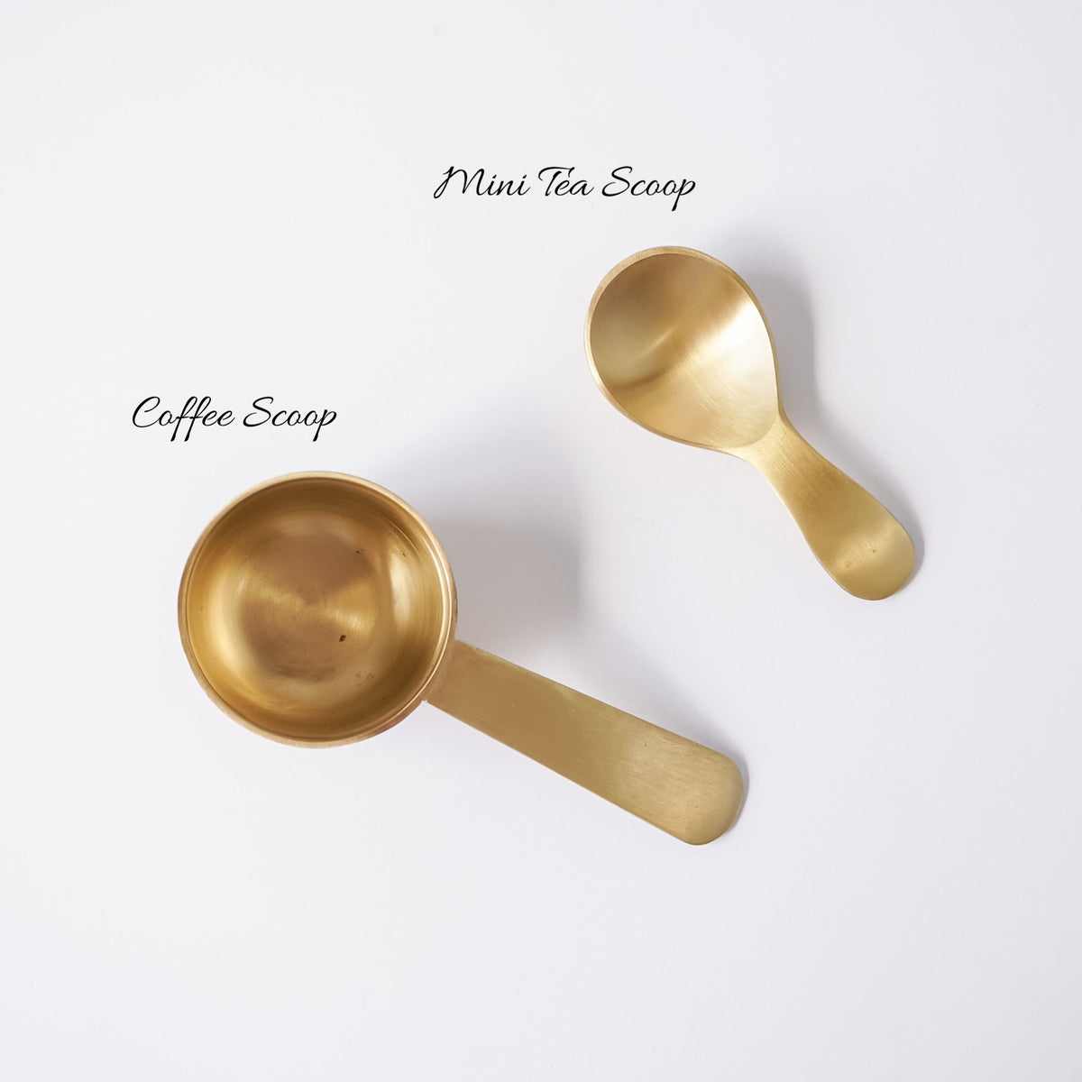 Two matte gold metal measuring cups, one larger scoop and one smaller spoon, arranged diagonally on a clean white background.