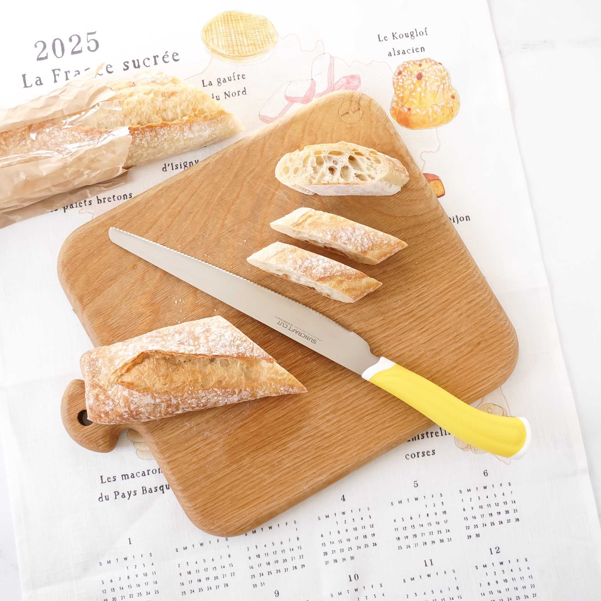 A bread knife with a yellow handle is placed on a wooden cutting board, which is surrounded by sliced bread and a calendar.