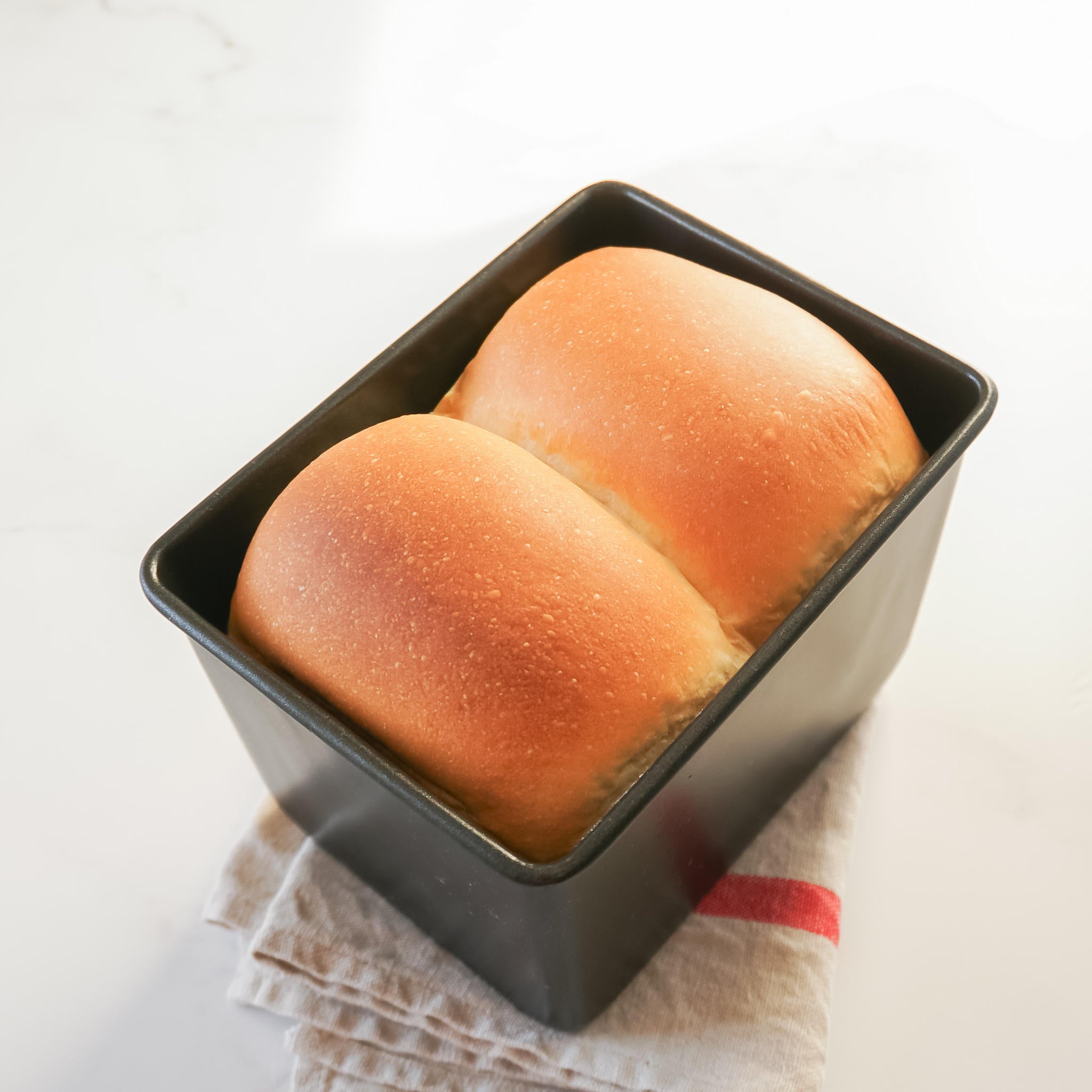 A black loaf pan filled with two loaves of freshly baked bread. The bread is golden brown and has a slightly crusty exterior.