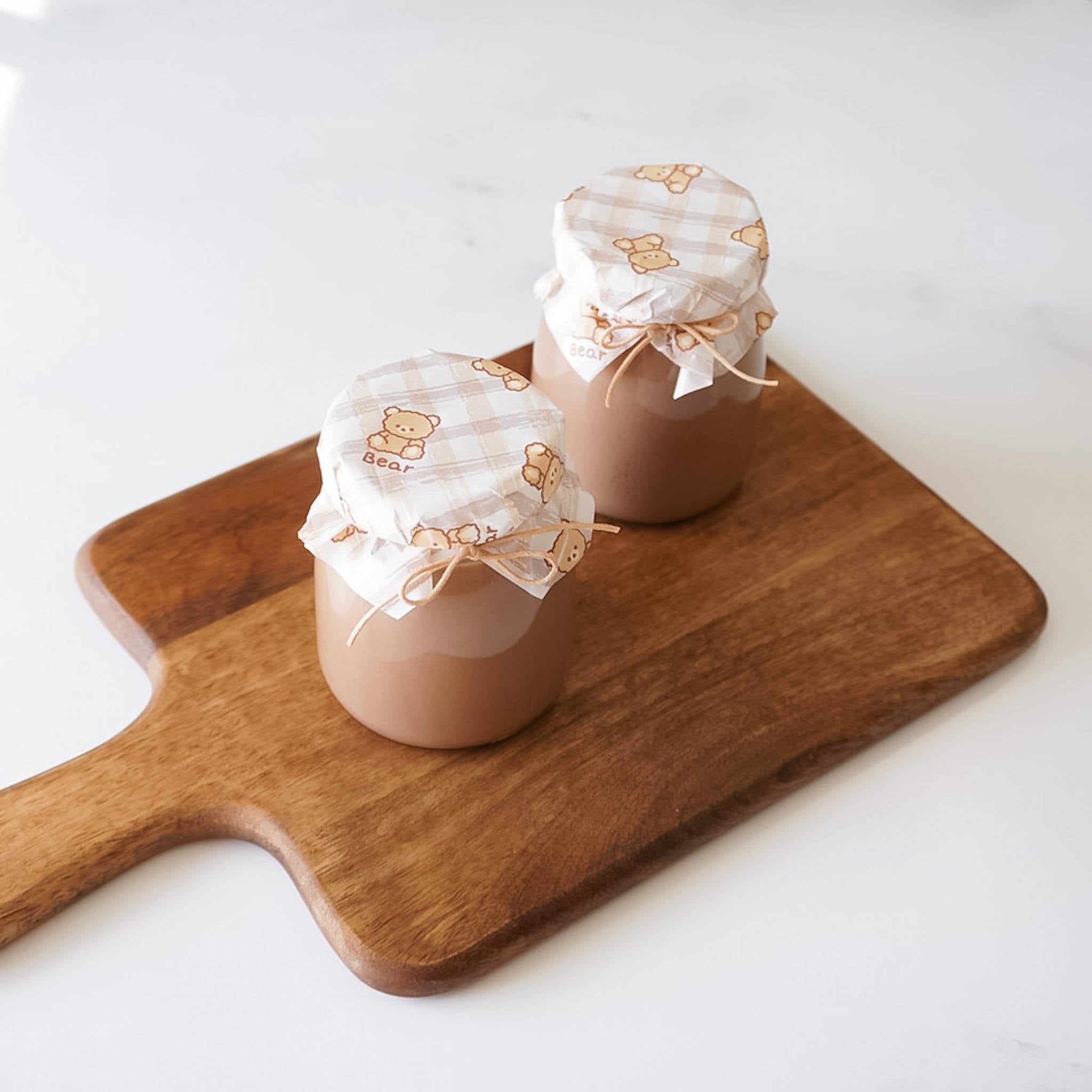 Two glass jars with brown liquid inside, topped with white deli paper covers with brown bear designs. The jars are on a brown wooden cutting board.