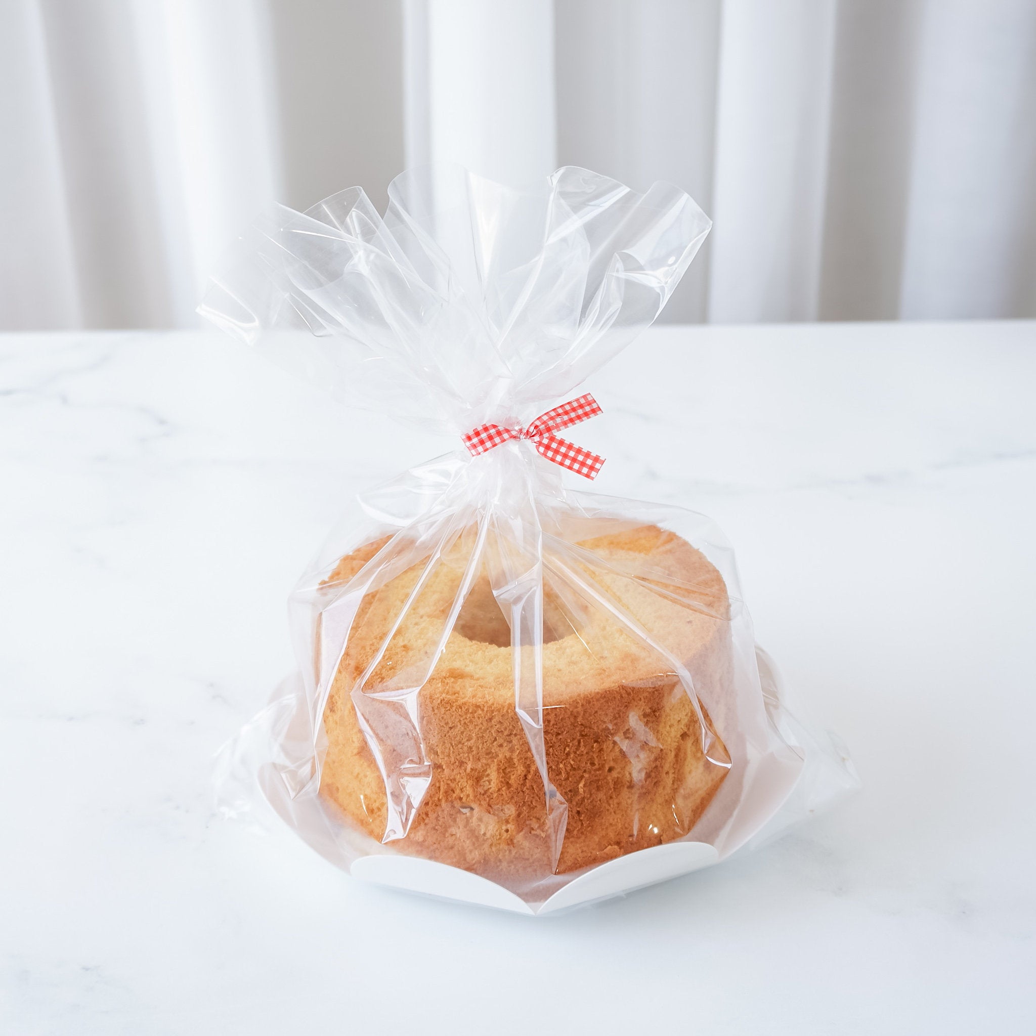 A cake packaged in a clear plastic bag with a flower-shaped cake board, tied at the top with a red and white checkered ribbon.