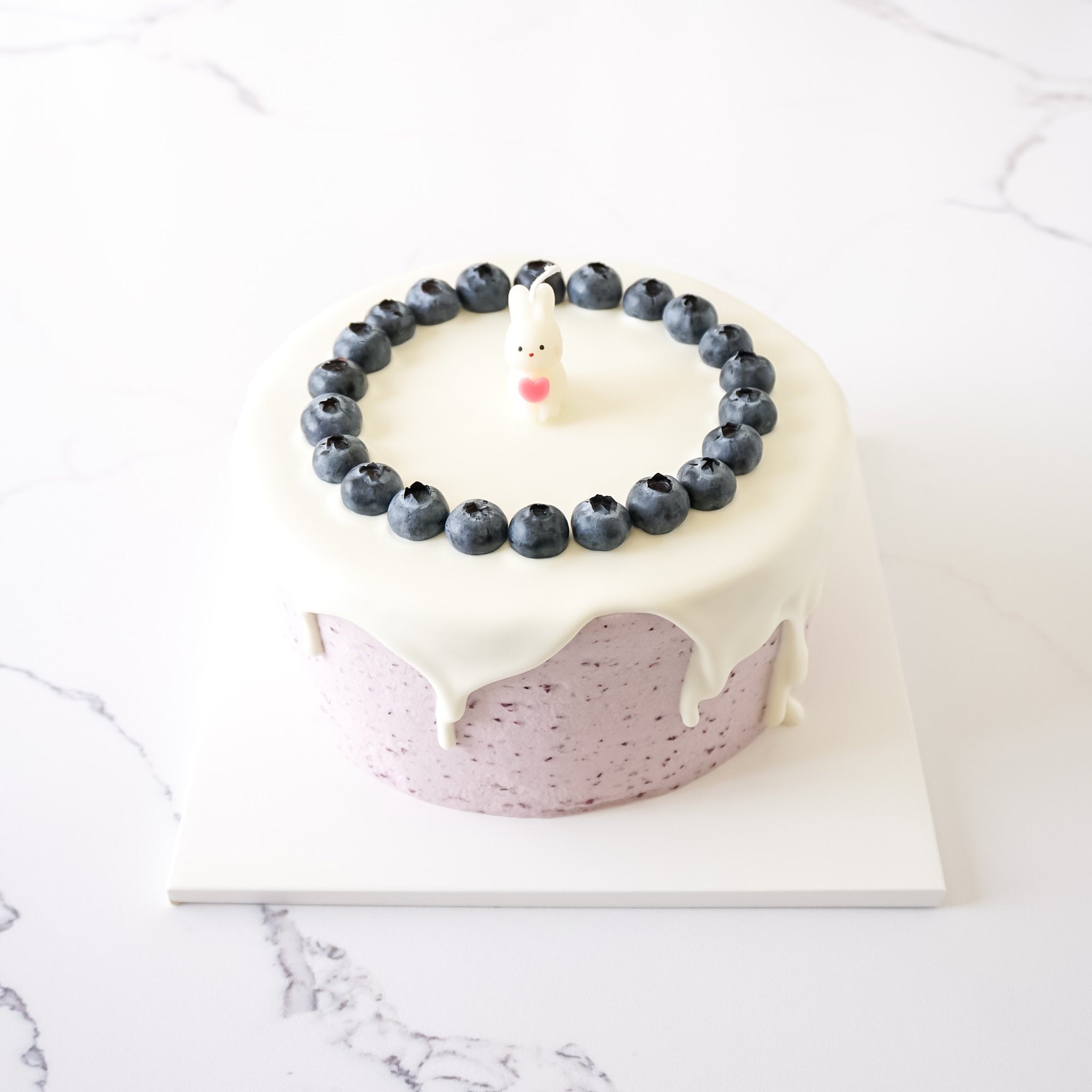 A cake candle with a pink heart-shaped bunny on top, surrounded by blueberries, placed on a white square plate with a marble background.