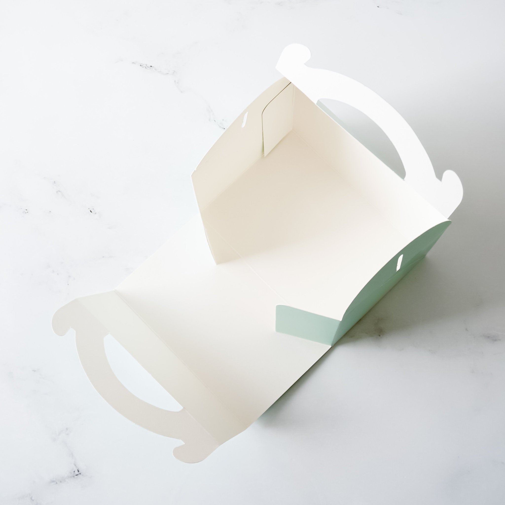 A white cardboard box with a green bottom. The box has two cut-out handles on the top and sides. The box is open and empty.