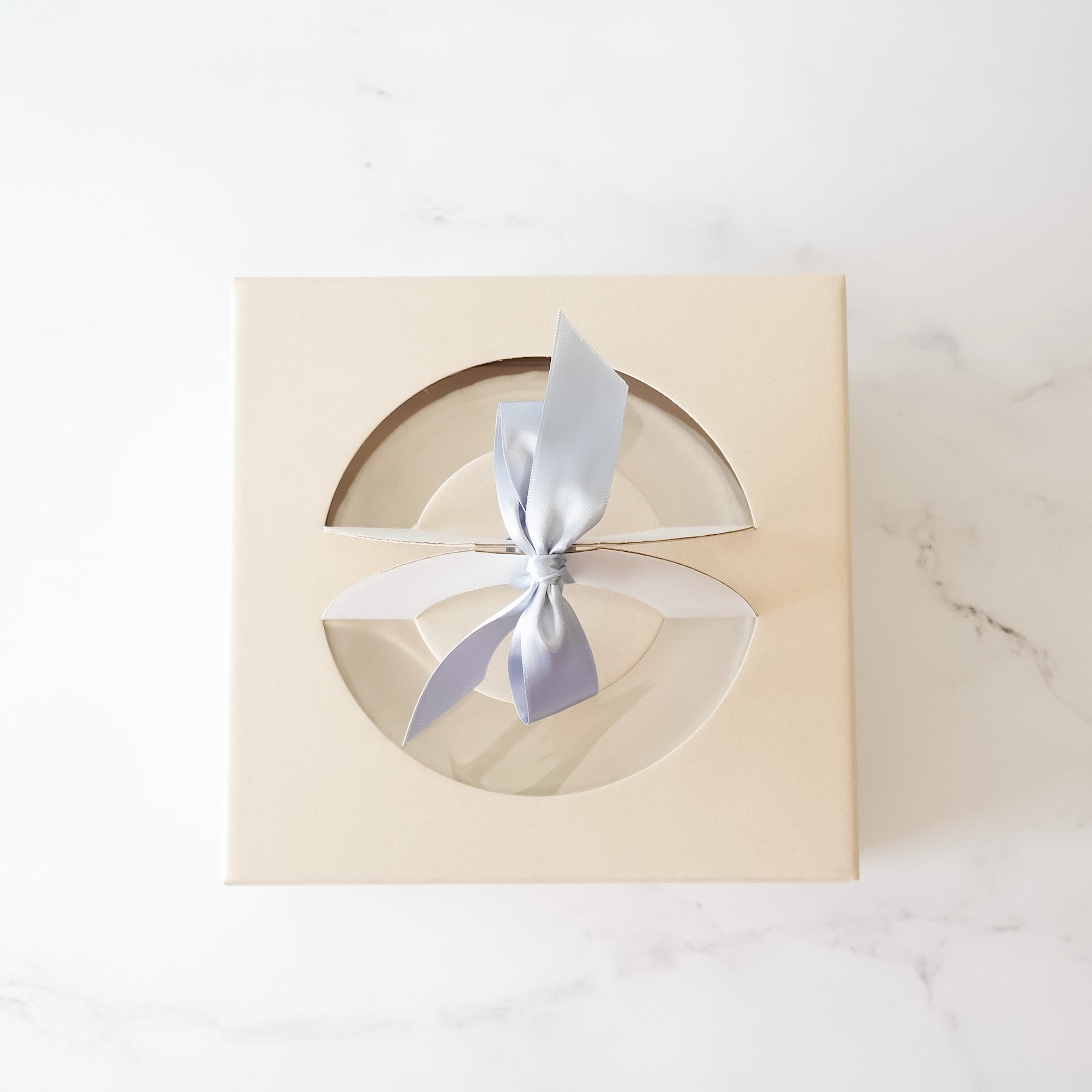 A beige cake box with a light blue ribbon tied in a bow. The box has a circular cutouts that reveal a white interior.