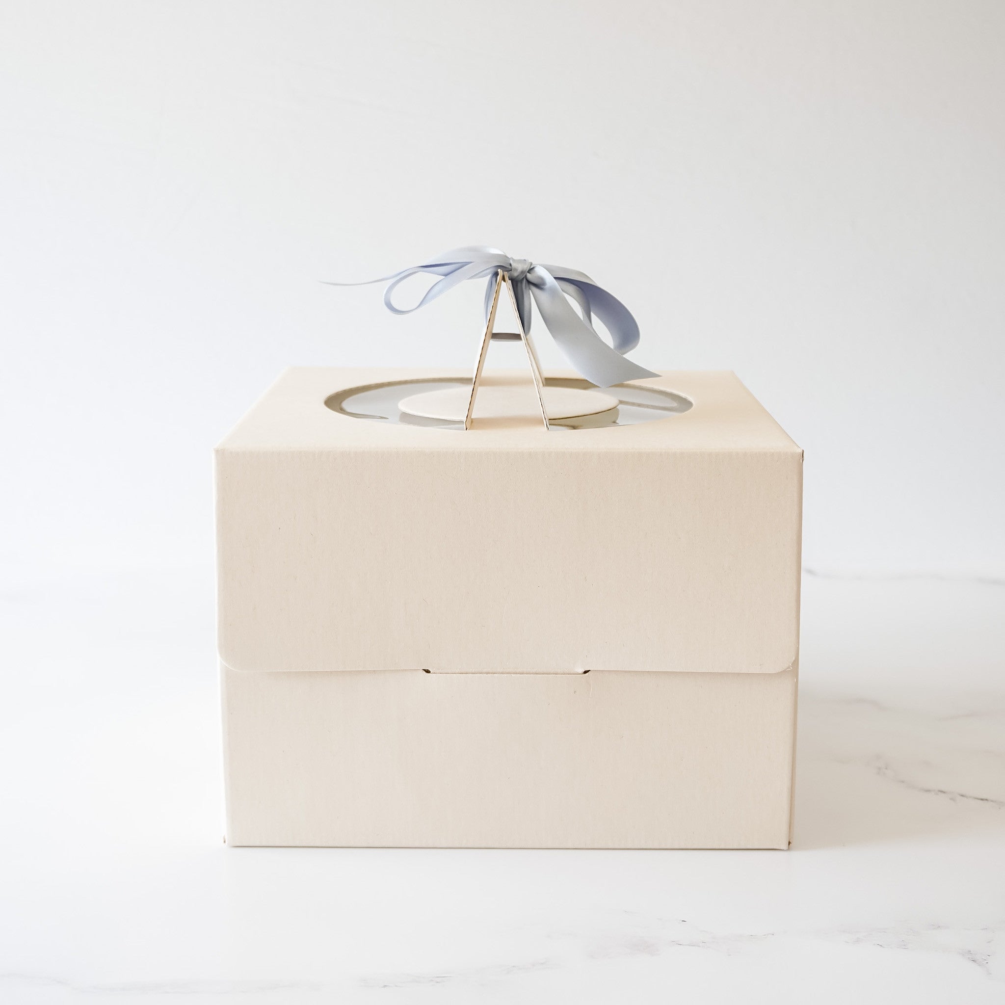 A beige cardboard cake box with a clear plastic window on top. A light blue ribbon is tied around the top of the box.