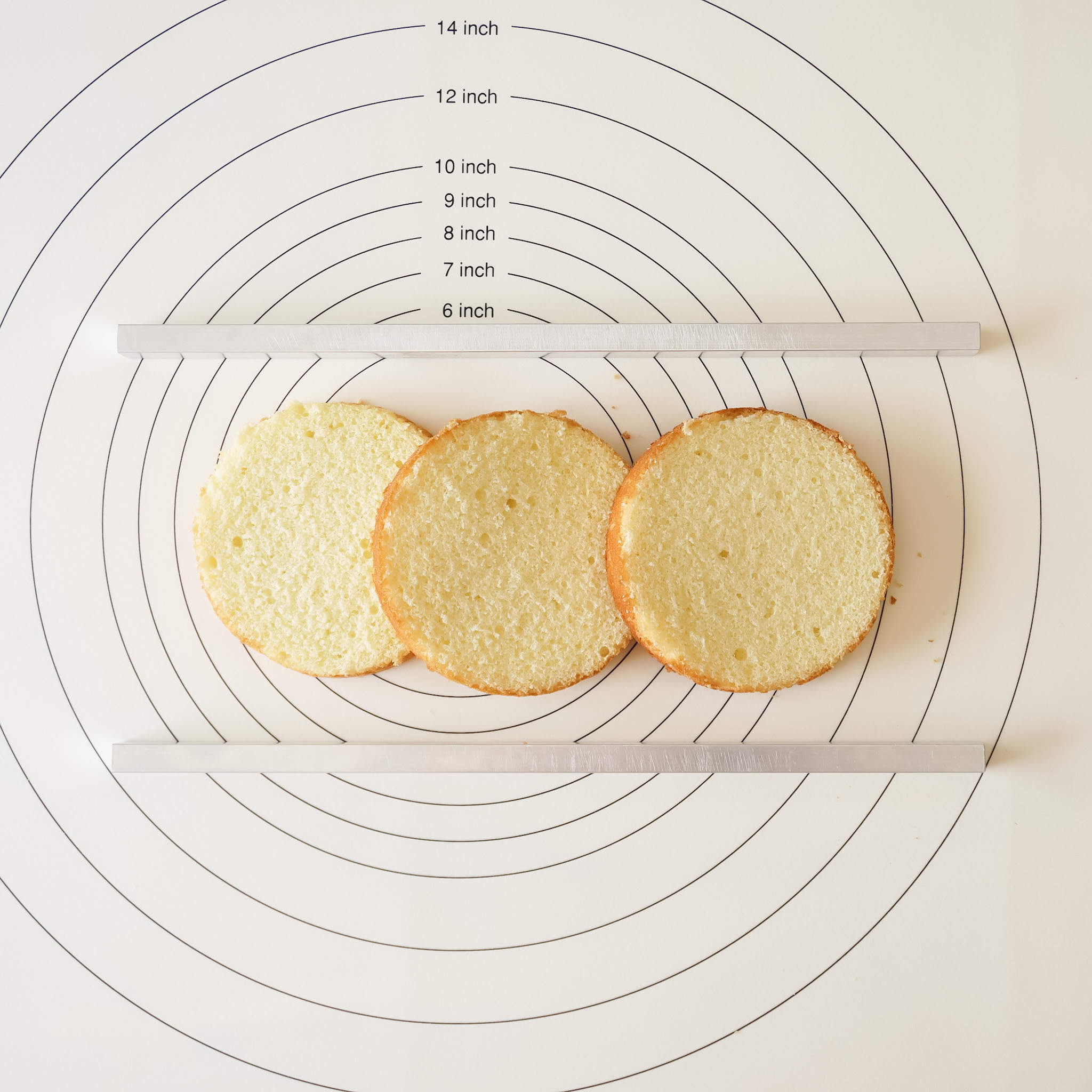 Two cake leveler tools placed horizontally on a surface with a silicone mat, alongside three layers of a cake displayed on top of the leveler.