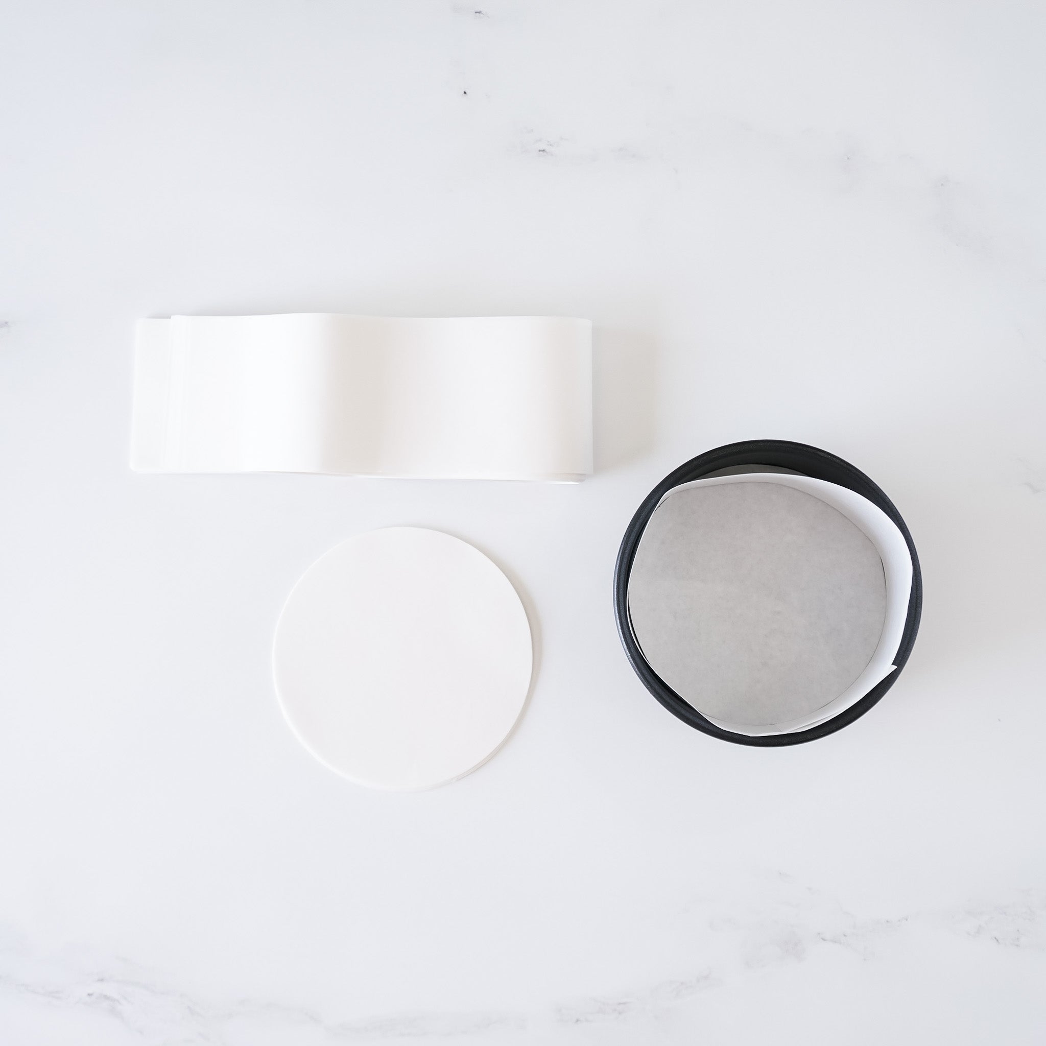 Precut white circle cake pan liners and side liners on a light background, with one side liner being black.