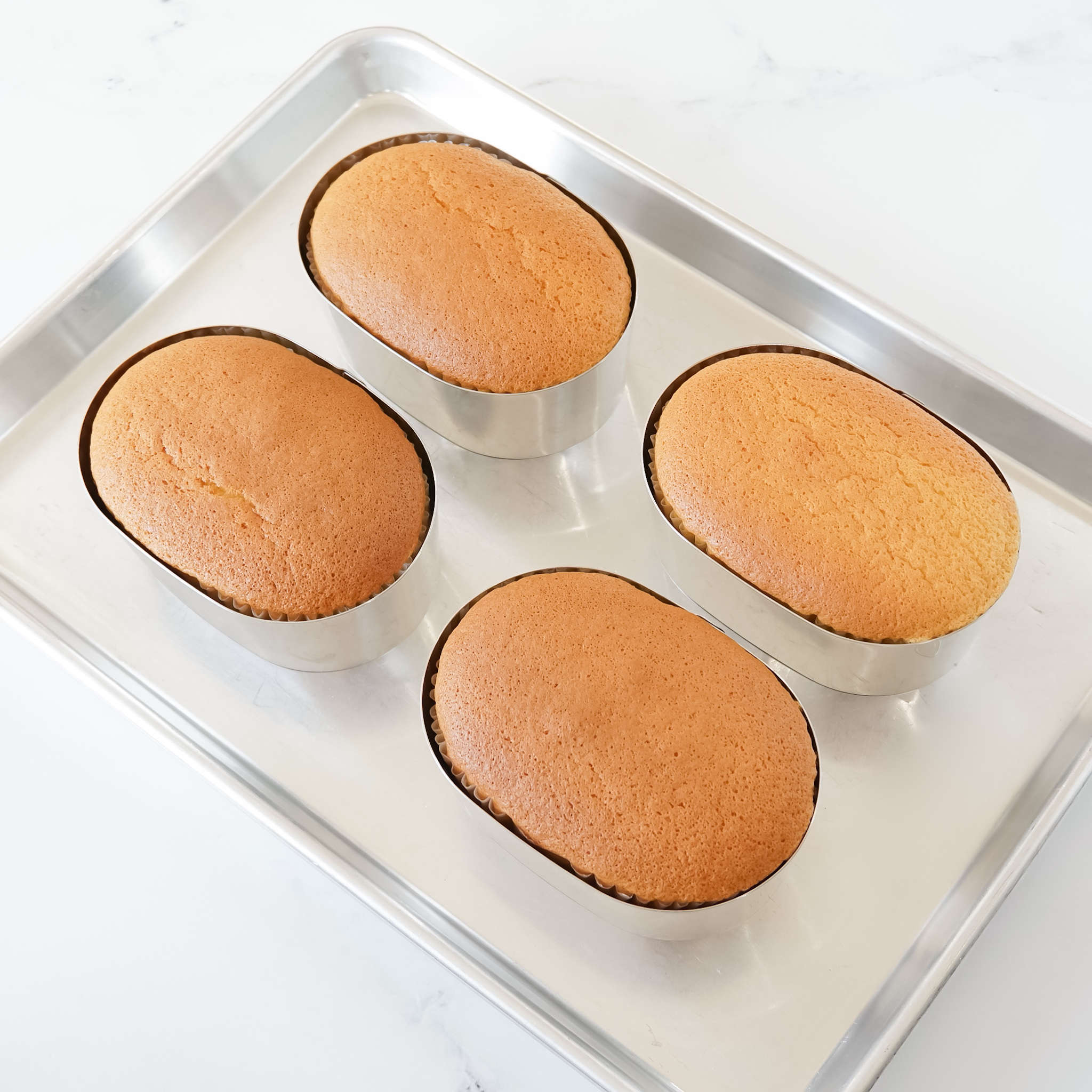 Four oval-shaped cake rings with a freshly baked castella cakes inside on a white marble surface.