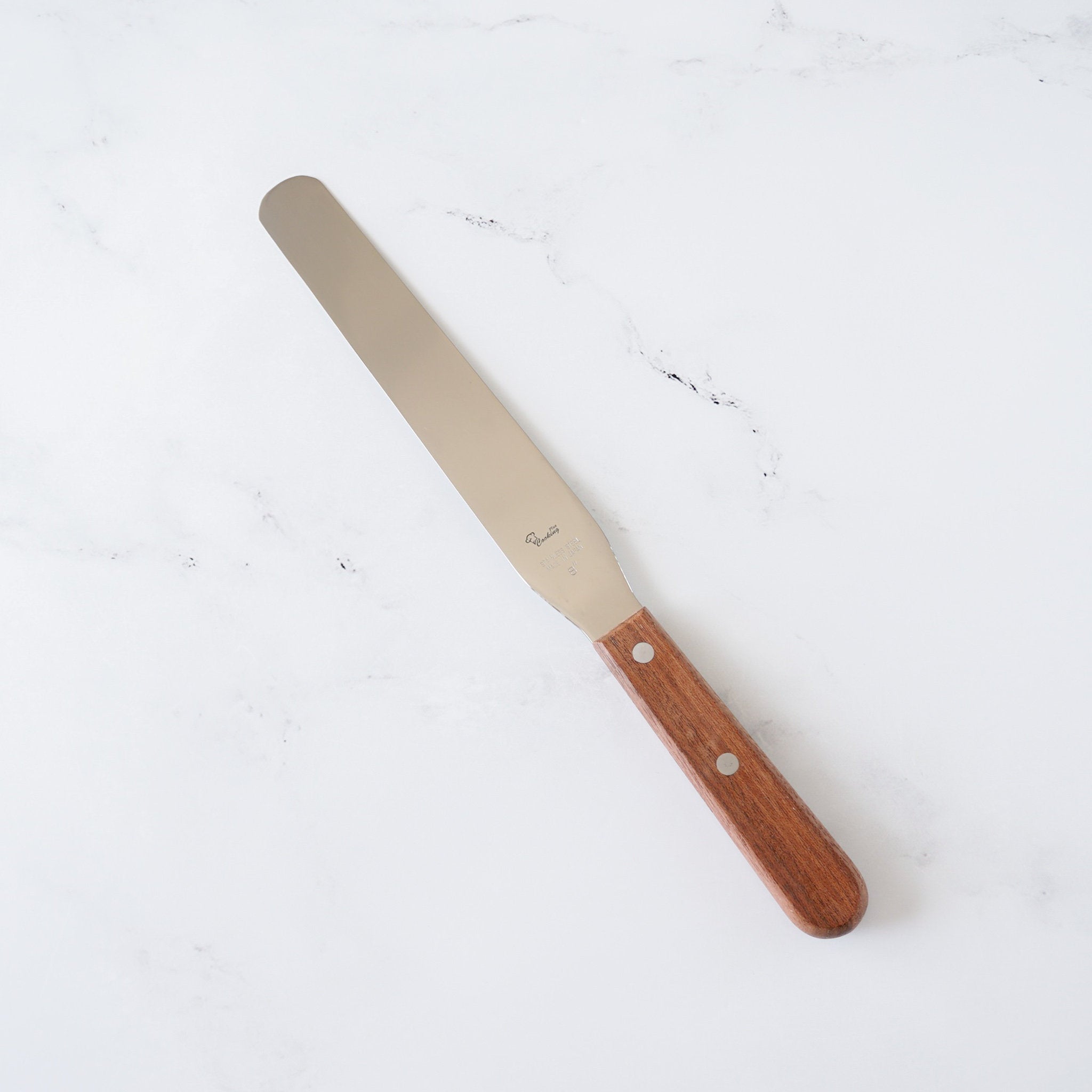 A long, silver spatula with a wooden handle. The handle is brown and has two silver rivets. The spatula is on a white marble surface.