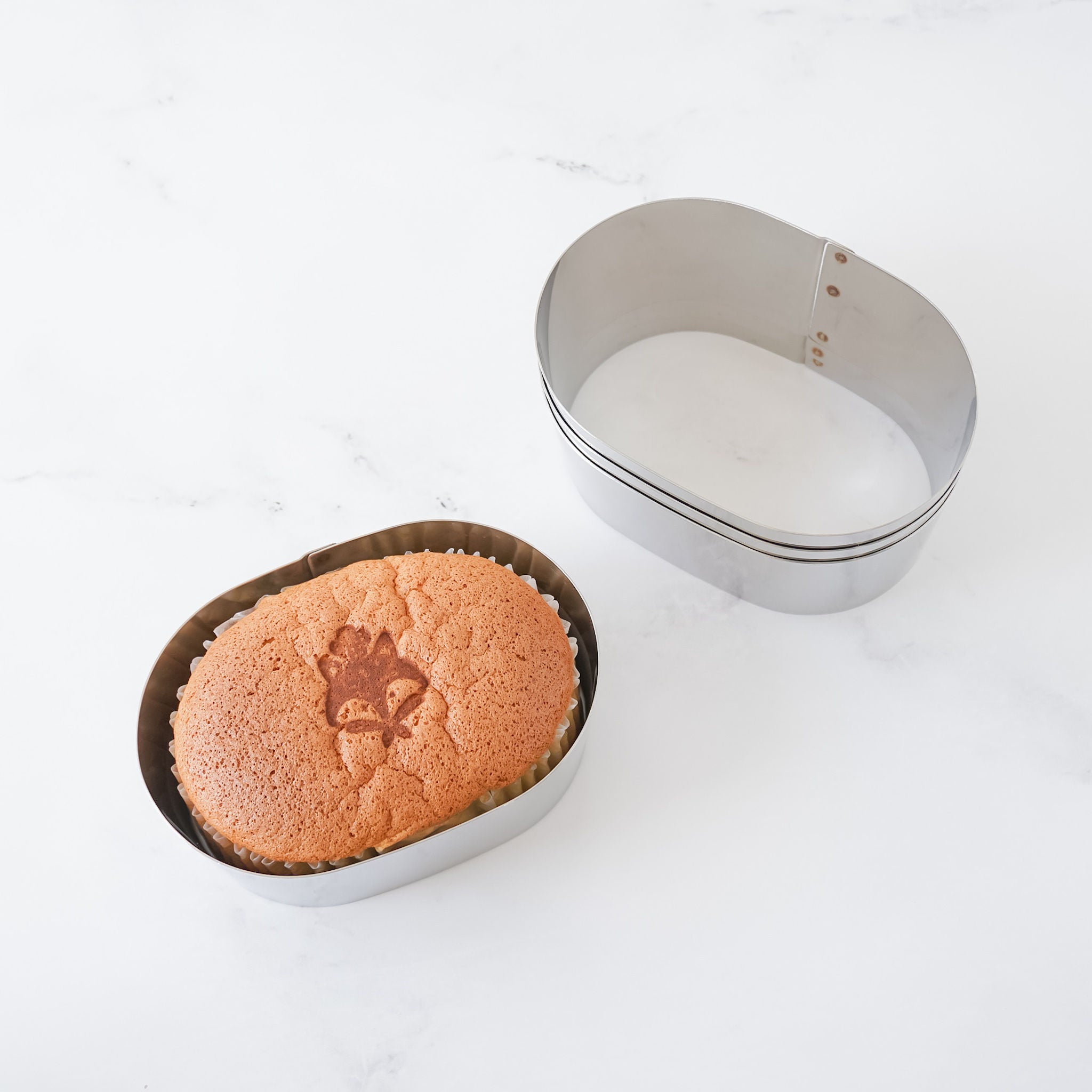 An oval-shaped cake ring with a freshly baked cake inside, alongside an empty ring, on a white marble surface.