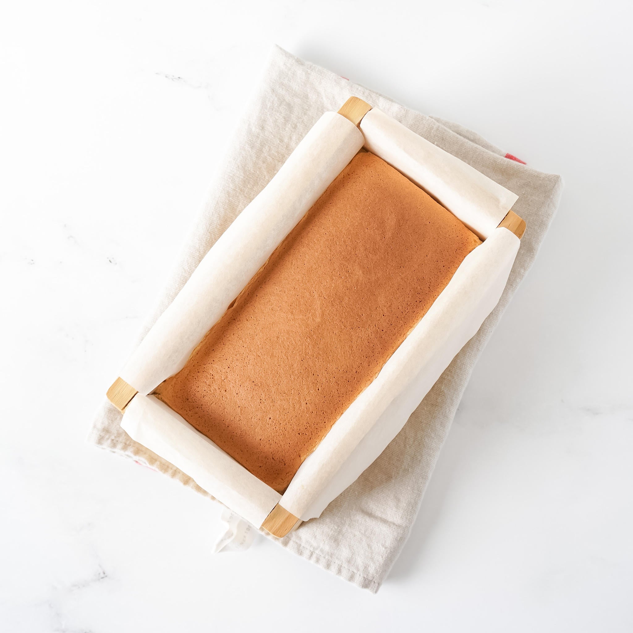 A freshly baked Castella cake in a wooden mold, which is placed on a kitchen cloth surface on a white background.