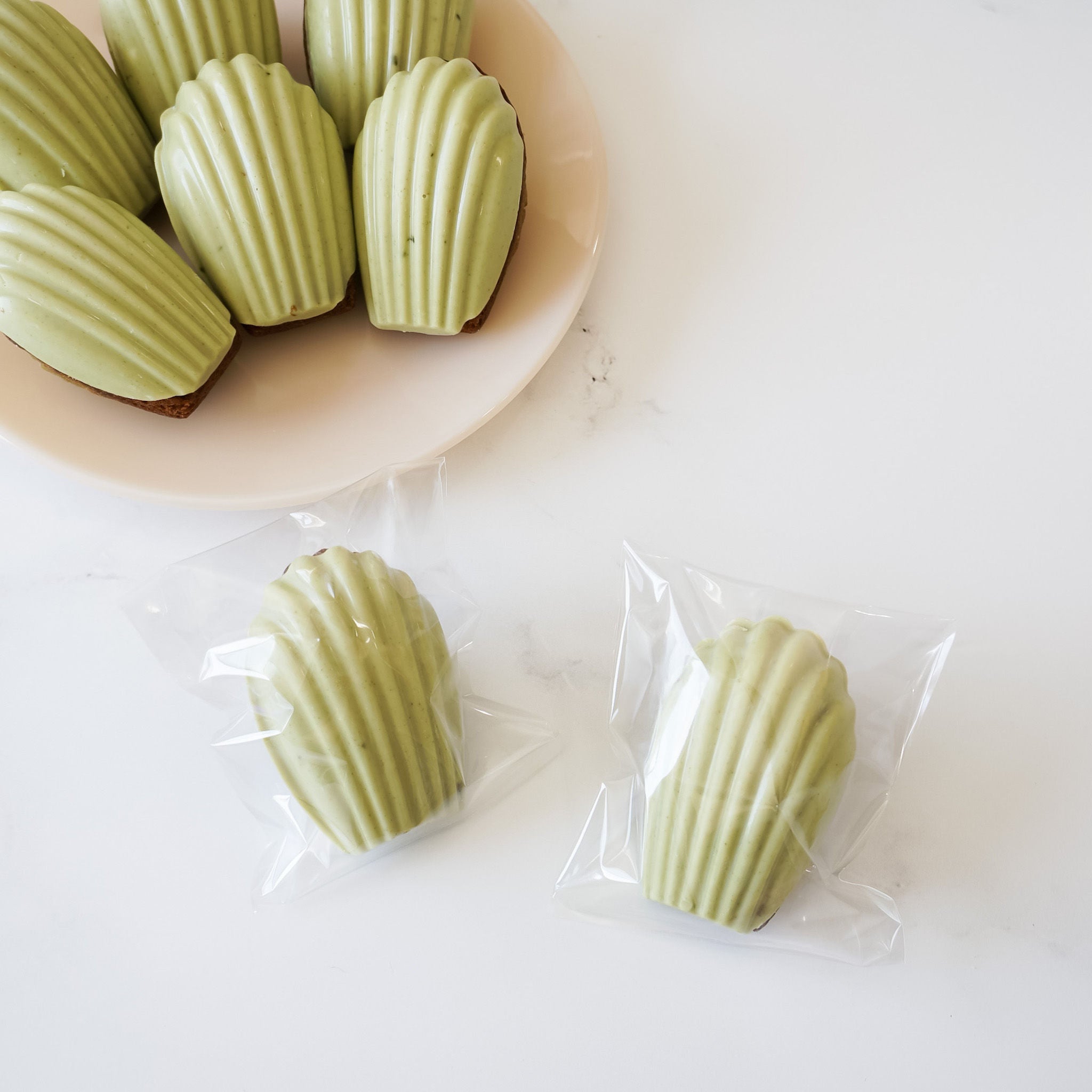 Two clear plastic bags each containing a single matcha madeleine. The madeleines are arranged on a white surface. There are six additional matcha madeleines on a white plate.