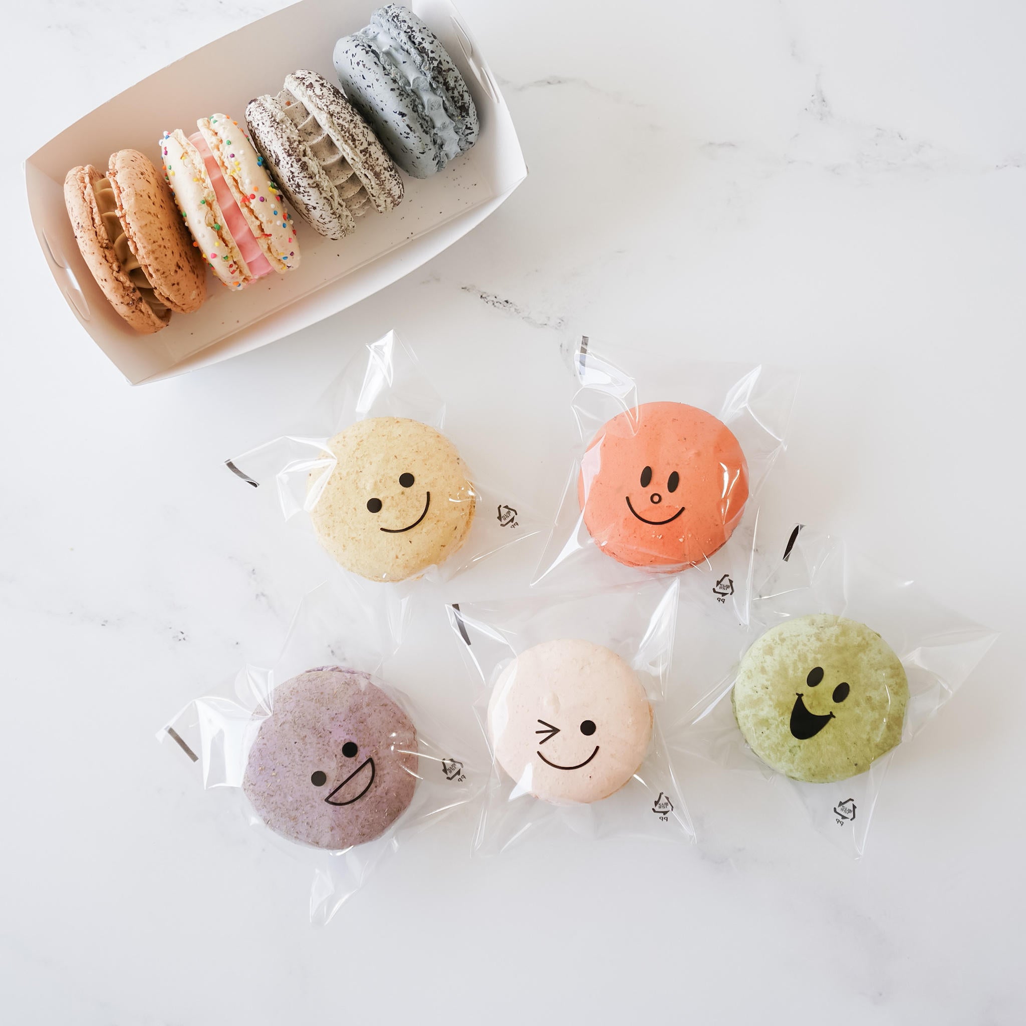 Six macarons in clear plastic bags with different smiley faces on them. The macarons are in the colors yellow, orange, purple, green, and white. The macarons are arranged in a row on a white surface.