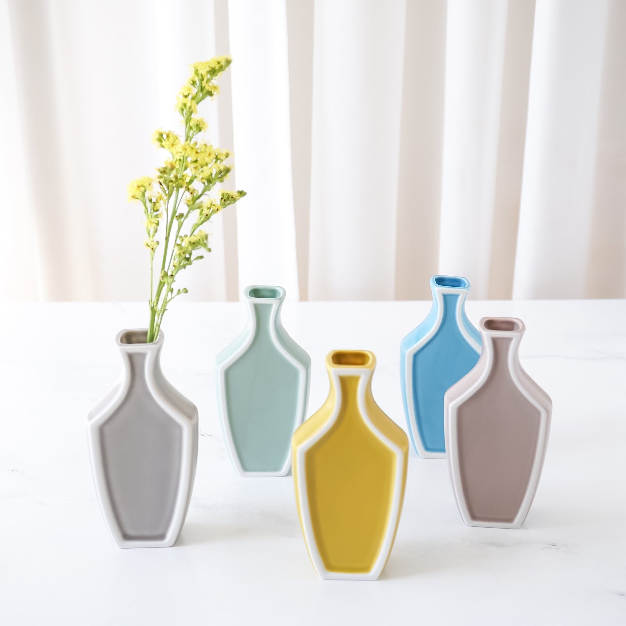 A collection of small ceramic vases in various colors, each with a white outline and a minimalist design, displayed on a white surface with a curtain background.