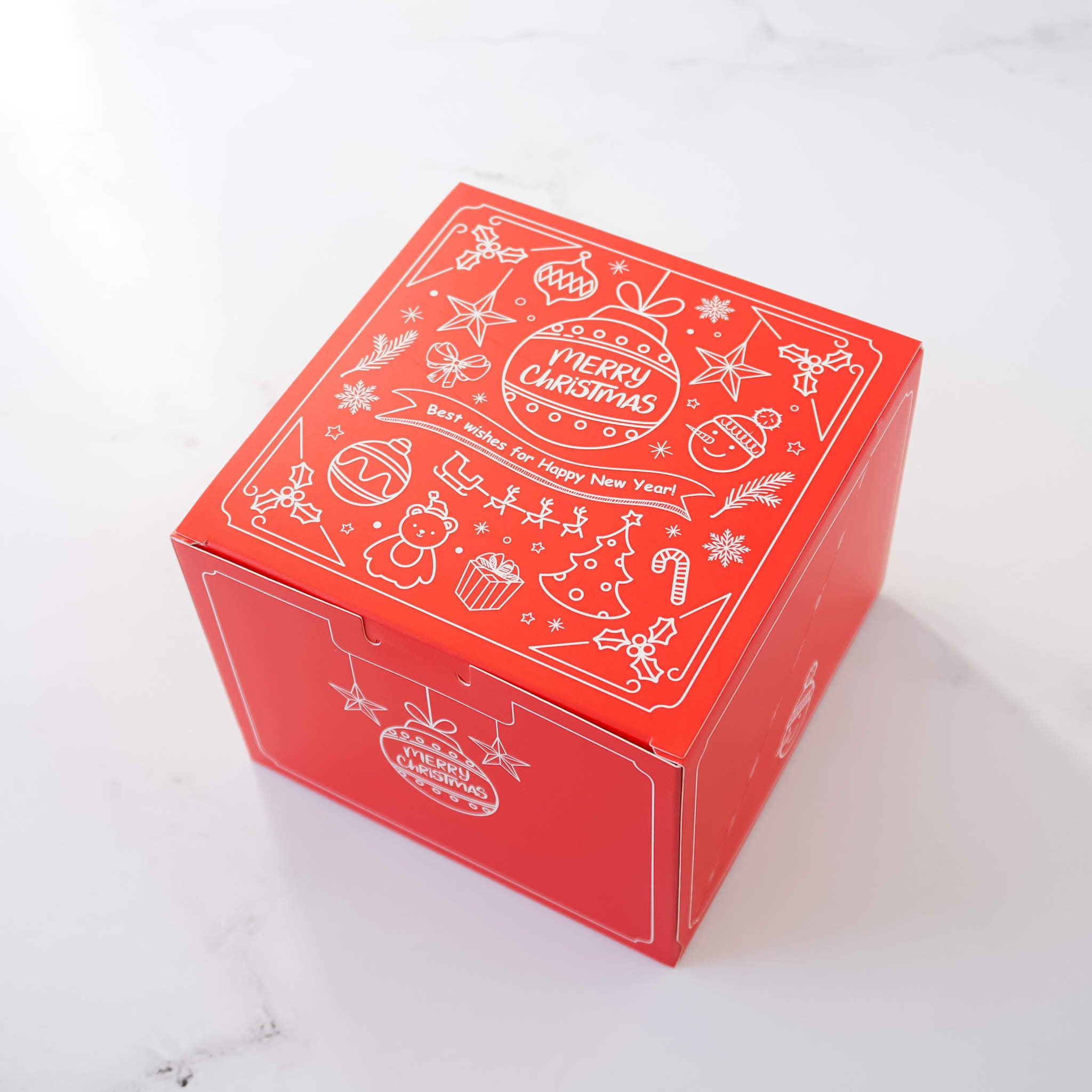 Red square Christmas gift box with white line art design. The box features a festive design with illustrations of Christmas ornaments, stars, candy canes, and a Christmas tree. The text on the box says &