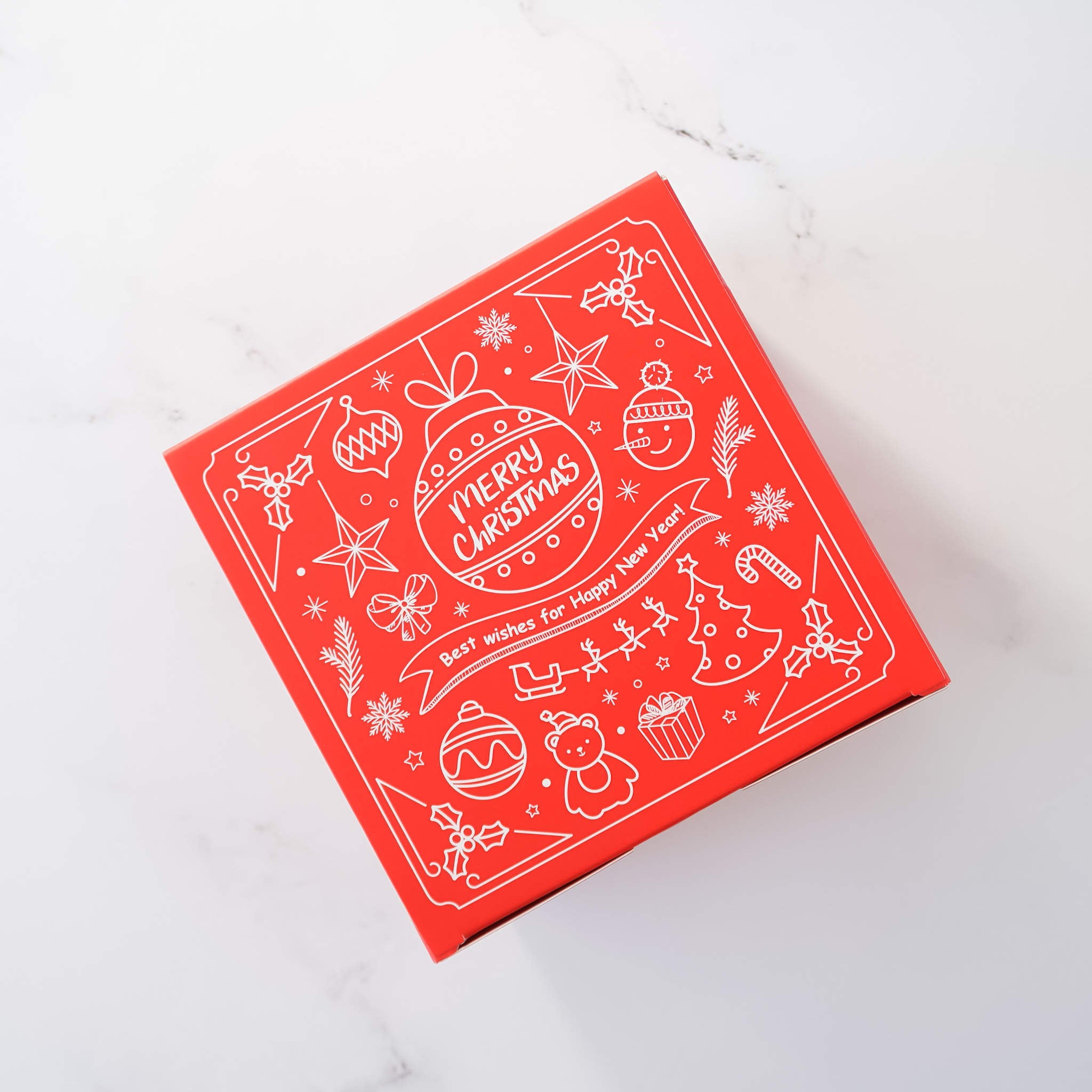 Red square Christmas gift box with a white line art design. The design includes illustrations of ornaments, stars, candy canes, snowmen, and a Christmas tree. The text on the box says &