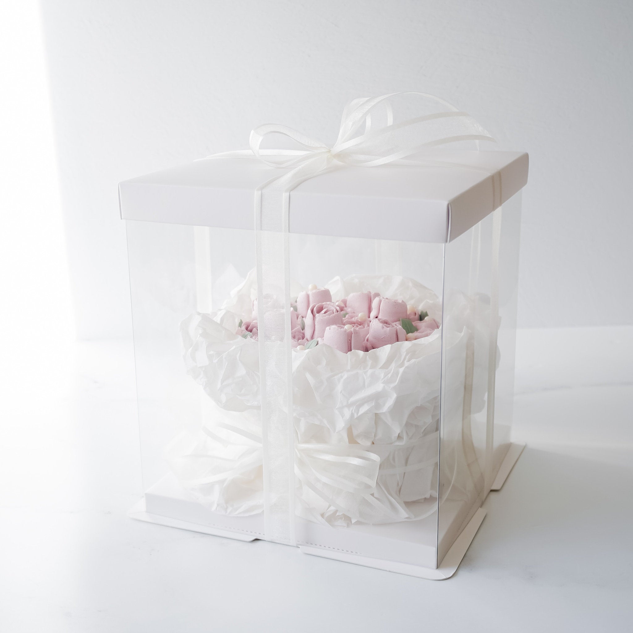 A white cake box with a clear plastic window containing a pink and white flower bouquet cake. The cake is wrapped in white tissue paper and tied with a white ribbon.