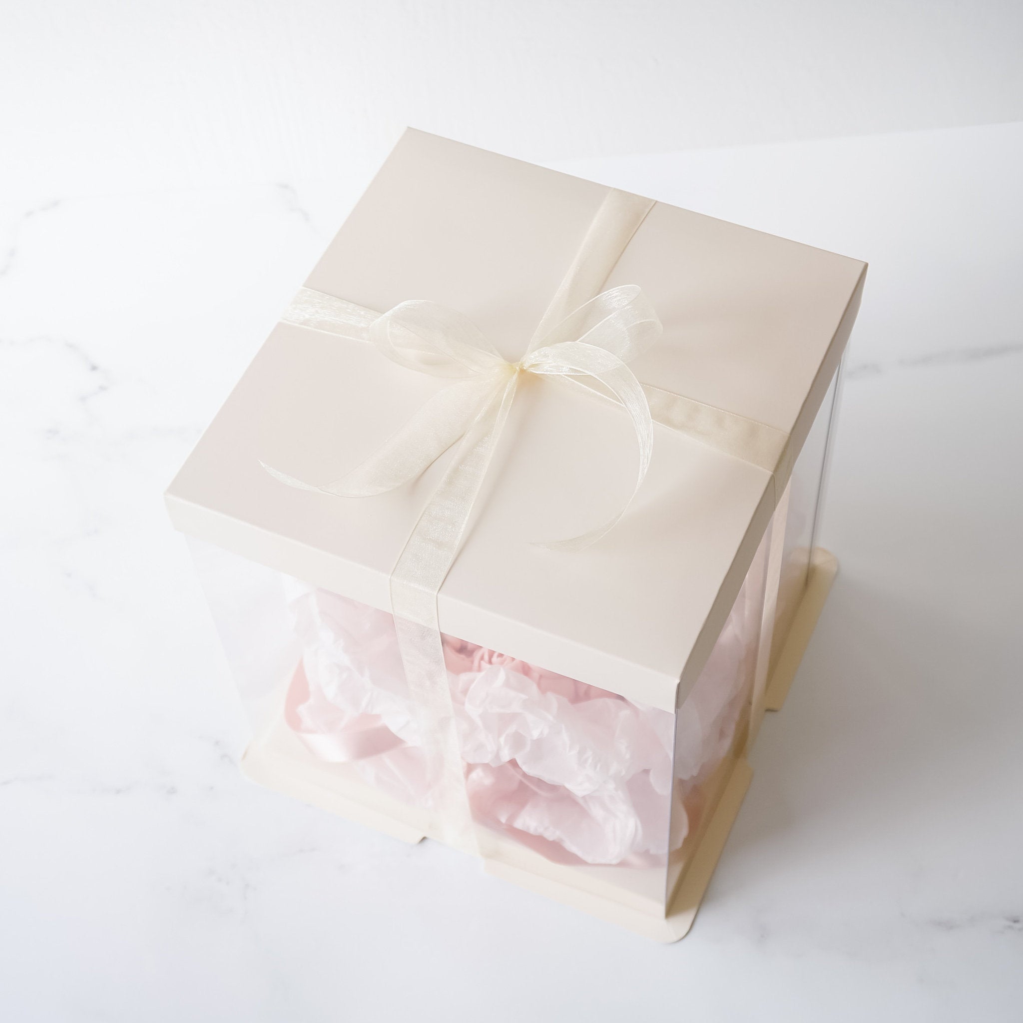 A clear plastic box with an ivory lid and a white satin ribbon tied in a bow. The box is filled with pink tissue paper.