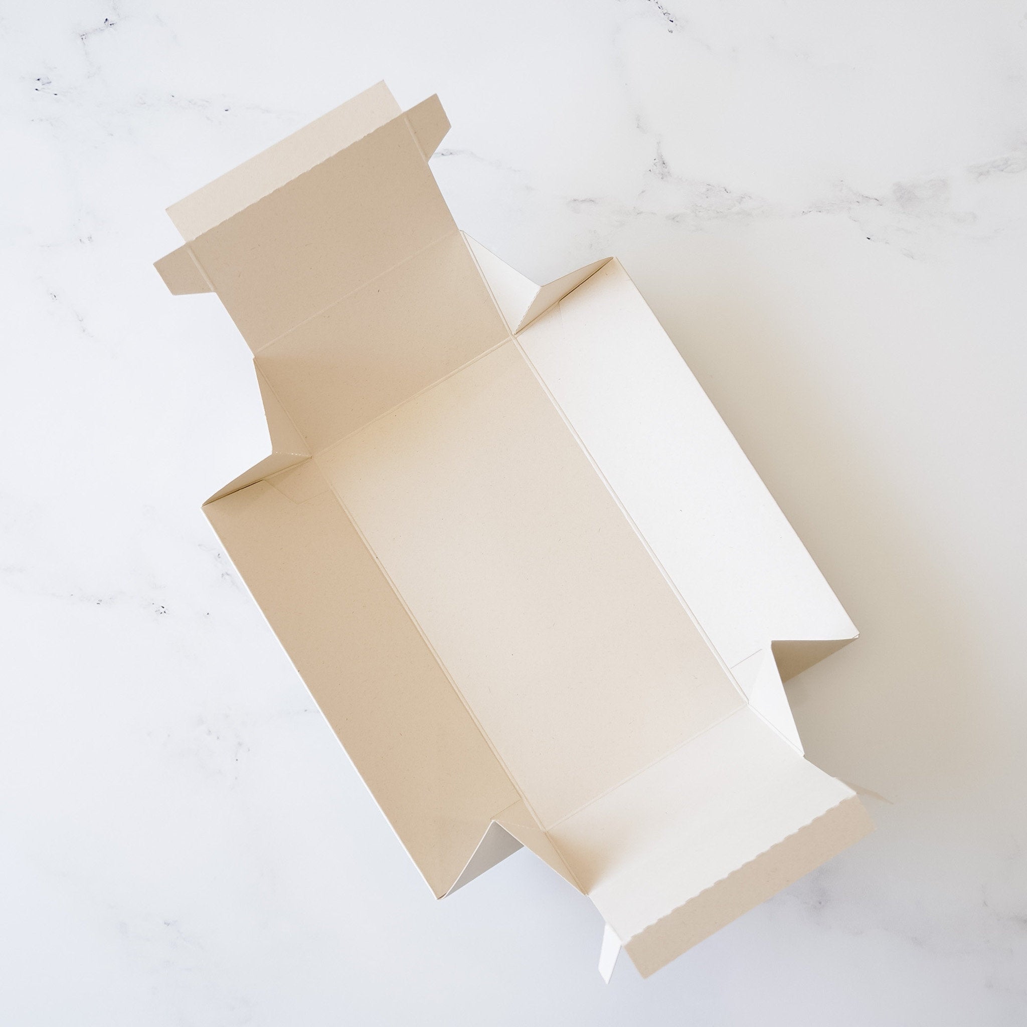 An empty, unfolded, rectangular cardboard box with a light beige color. The box has a flap on the top and two flaps on the sides. The box is lying flat on a white surface.