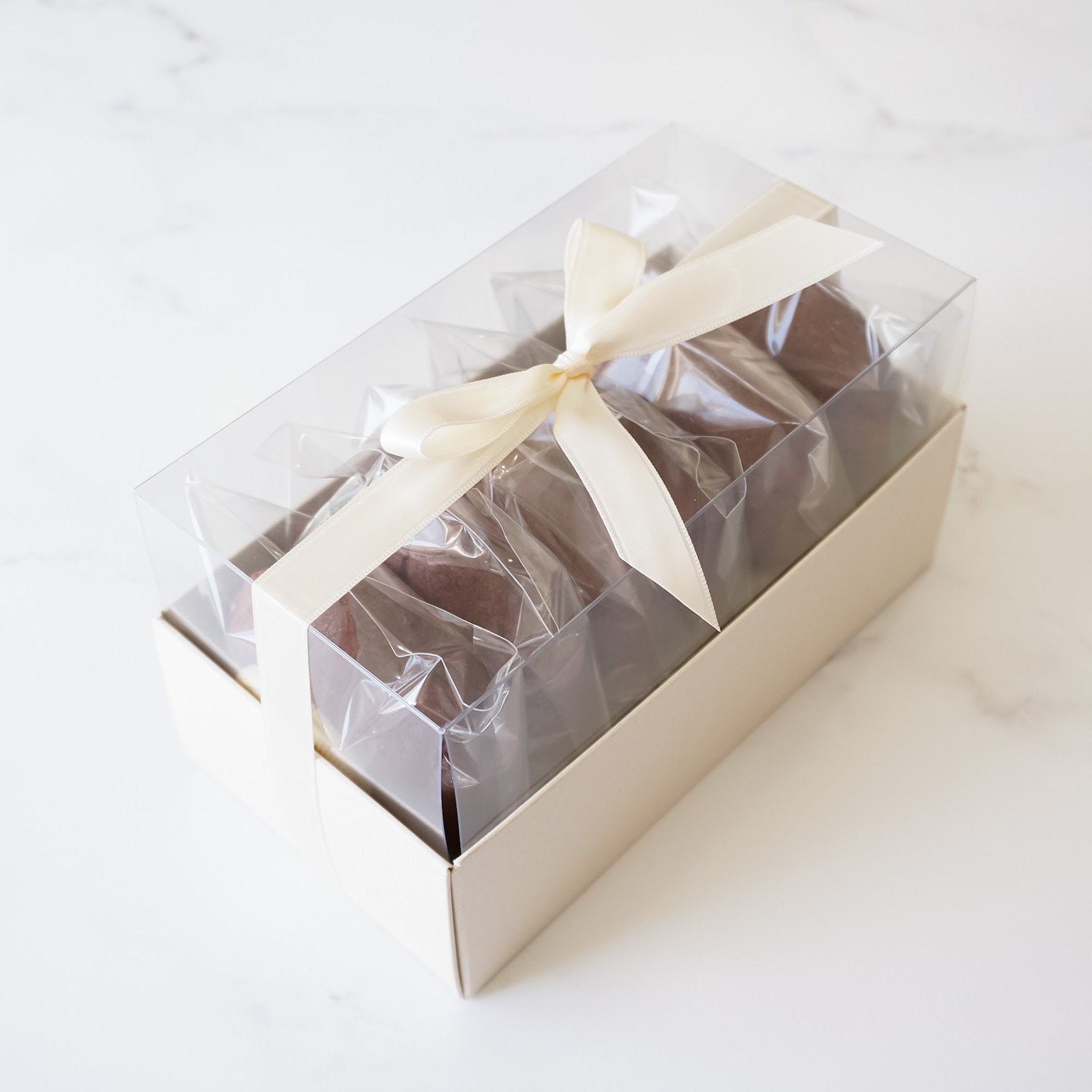 A beige cookie box with a clear lid, containing cookies wrapped in individual clear plastic packaging, tied with a satin bow.