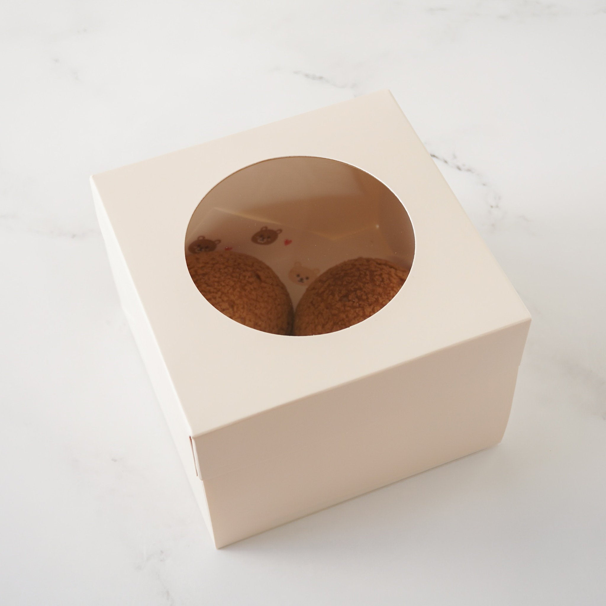 An ivory cardboard box with a round window. Two brown pastries are visible through the window.