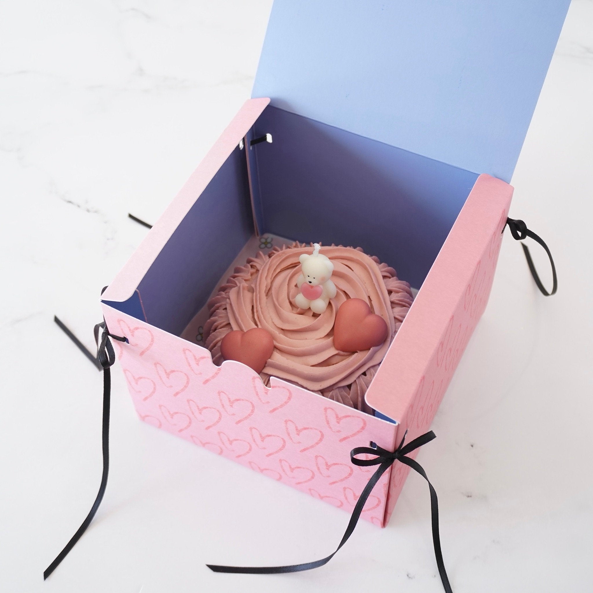 A pink box with a heart pattern and black ribbon handles. The box is open and contains a pink cake with a white teddy bear candle on top. The cake is decorated with pink frosting and two heart-shaped chocolates.