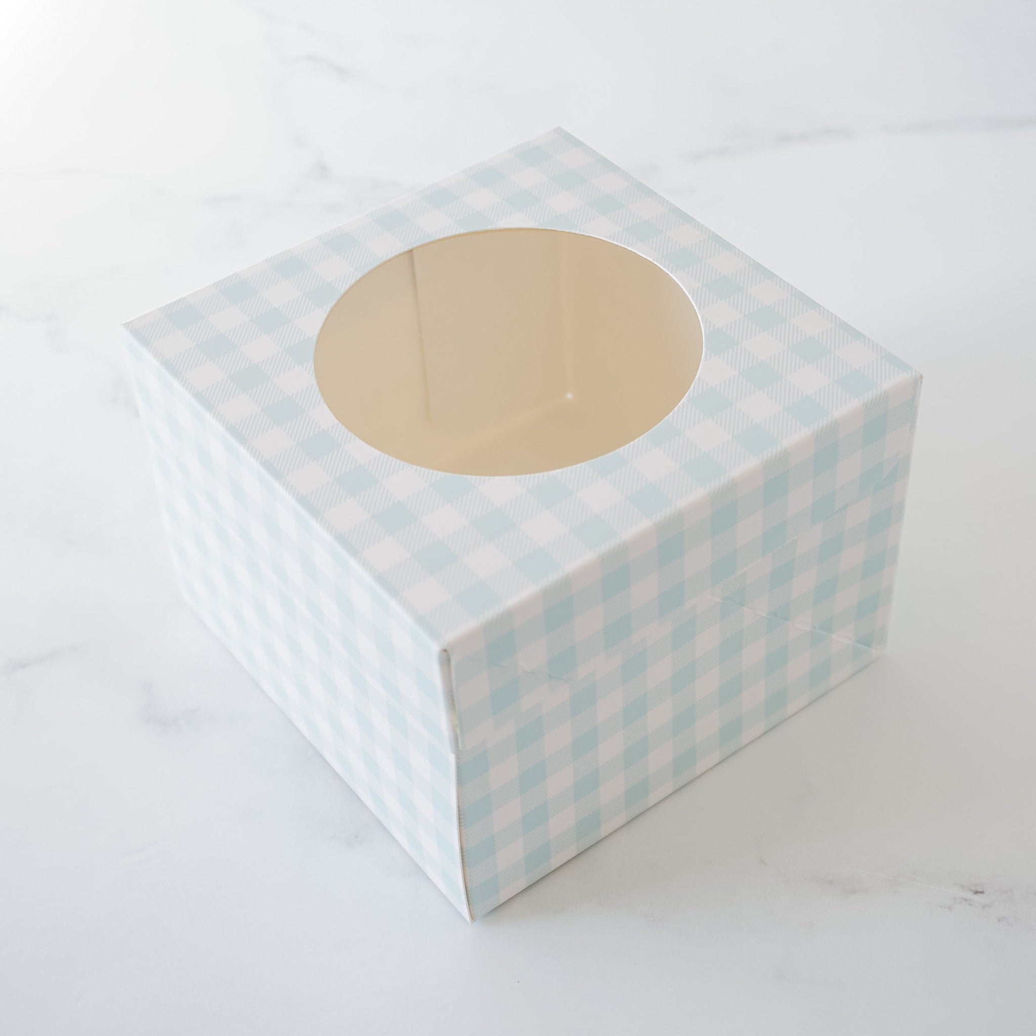 A square, light blue and white gingham patterned gift box with a round window in the center. The box is empty.