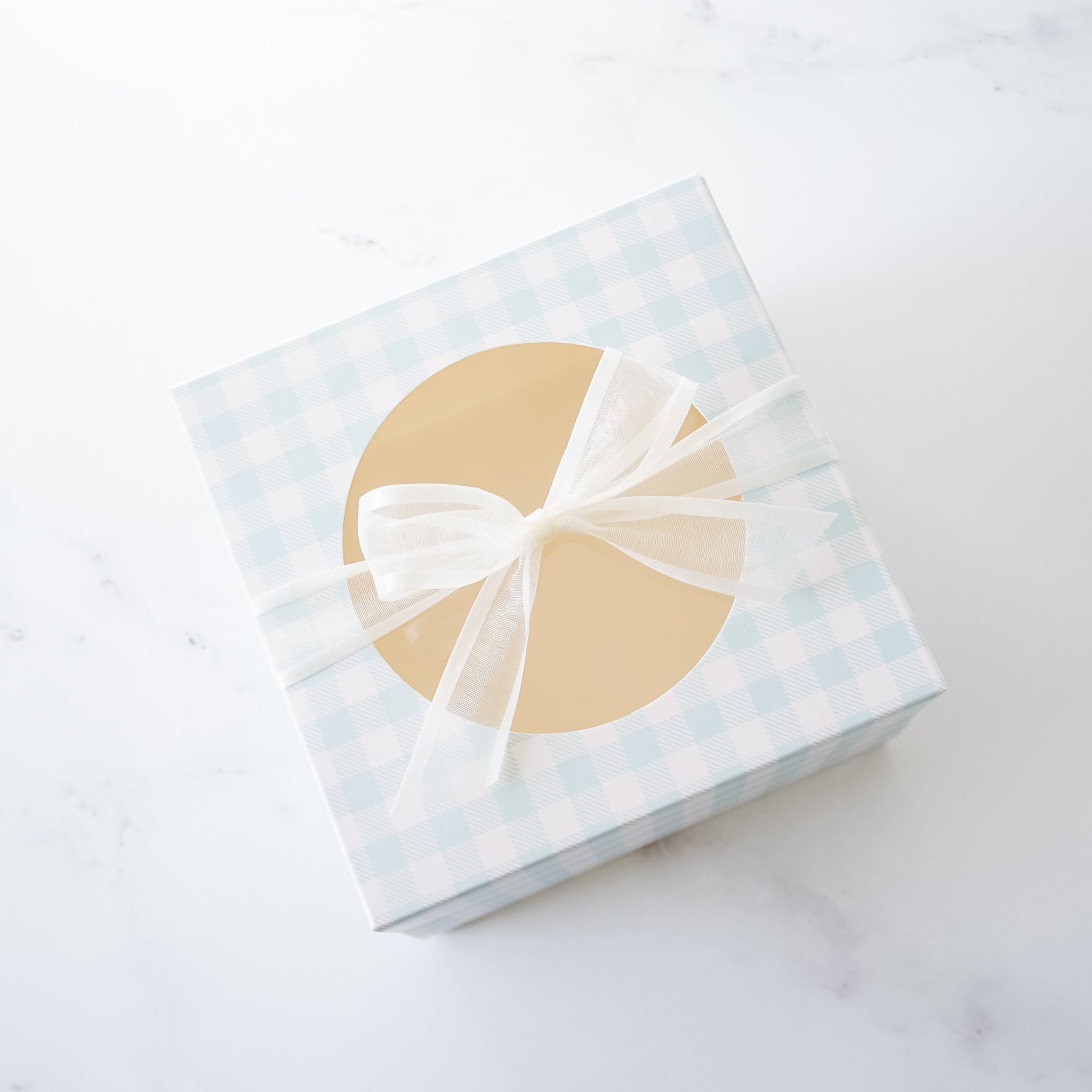 A closed Blue Gingham Cookie Box with a round window on top, decorated with a satin bow, on a marble surface.