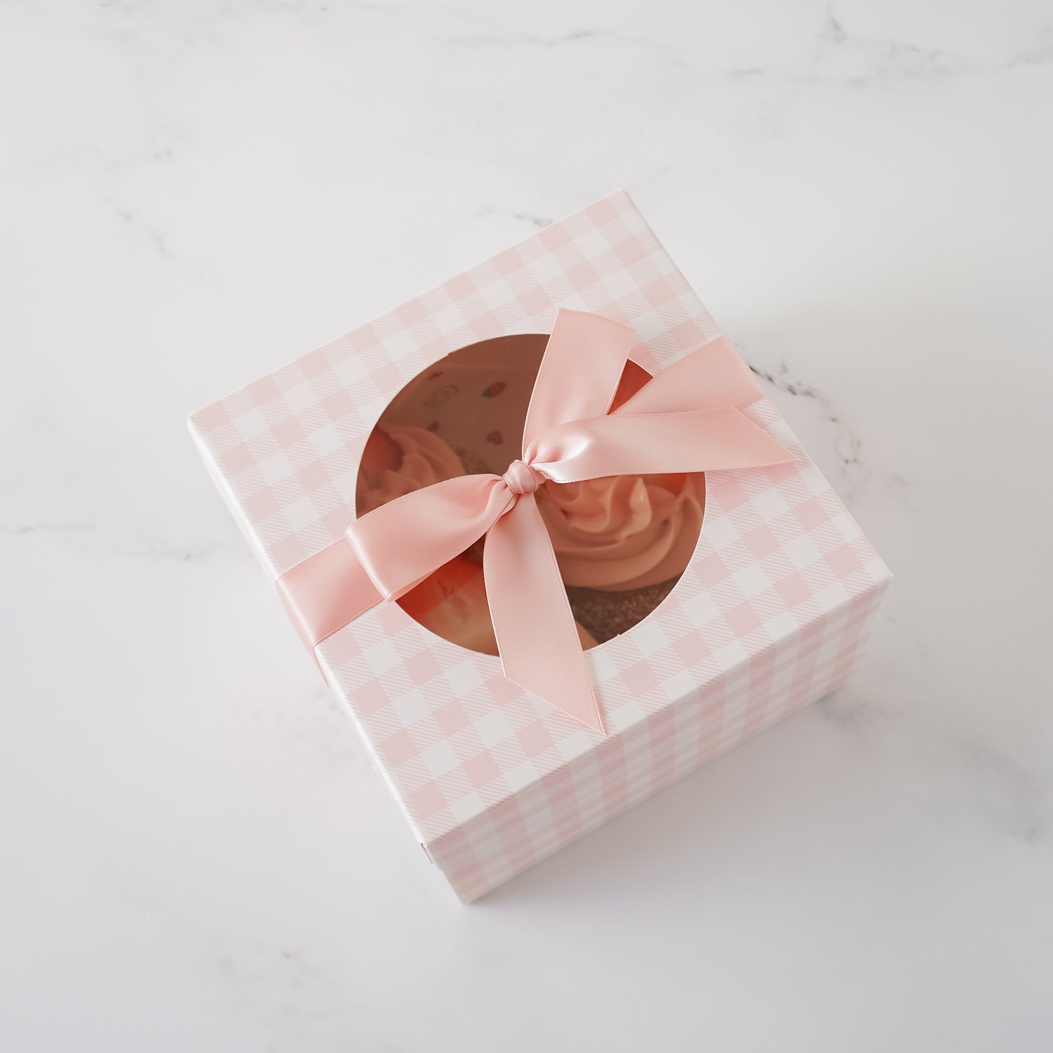 A pink gingham cookie box with a round window, tied with a pink ribbon, on a marble surface.