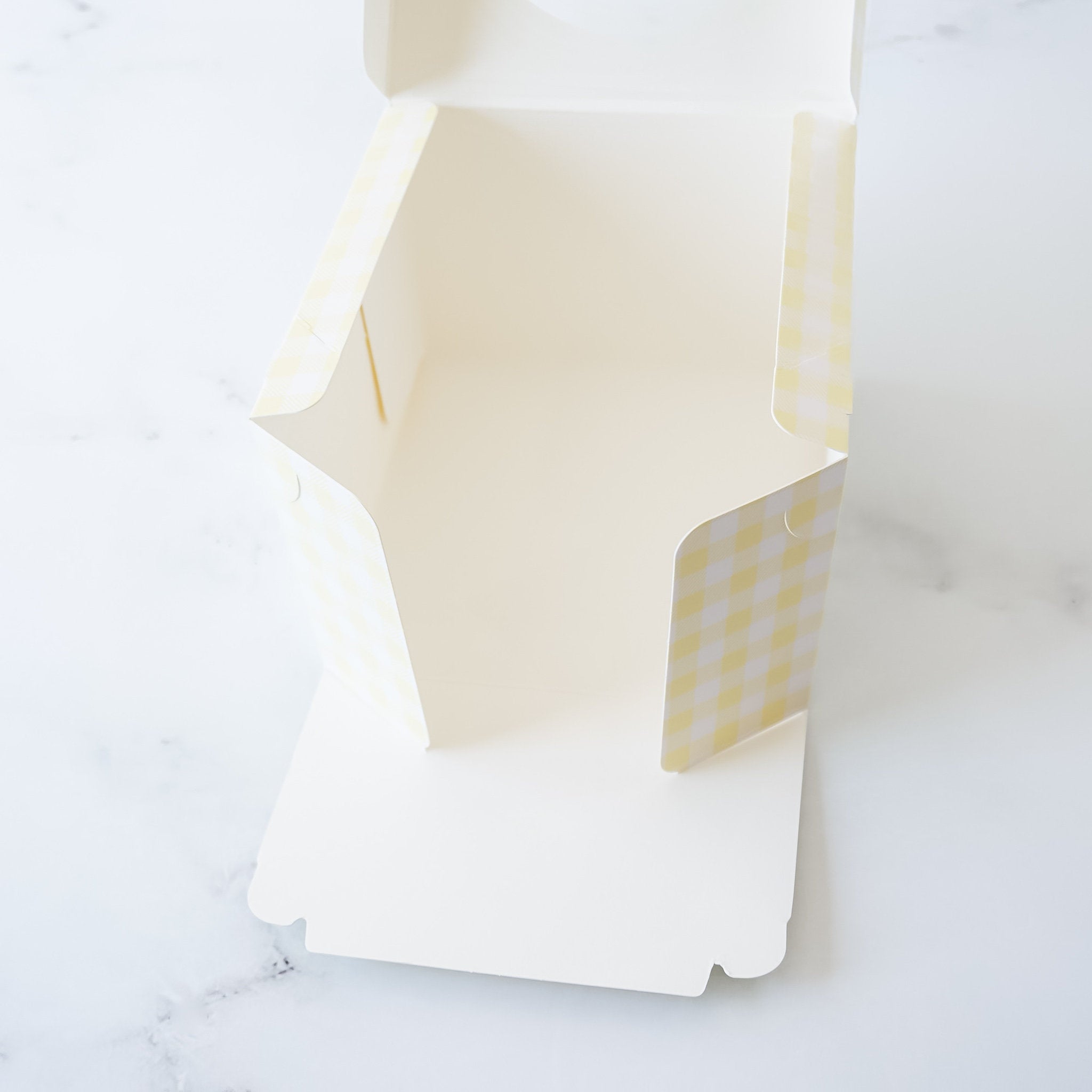 An open, empty, white cardboard box with a yellow and white checkered pattern on the inside. The box is designed for packaging baked goods.