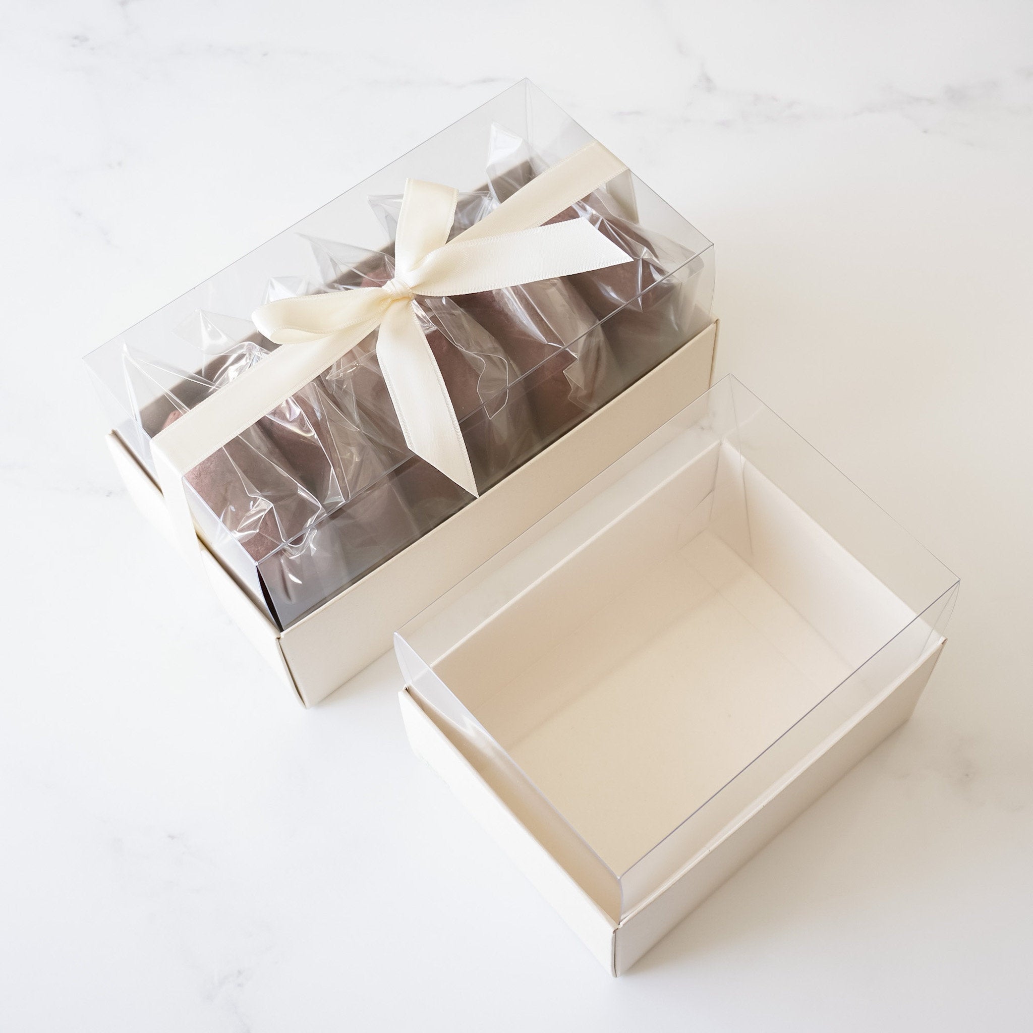 Two light beige boxes with clear lids. The box on the left has a white ribbon tied around it and contains several brown chocolate candies. The box on the right is empty.