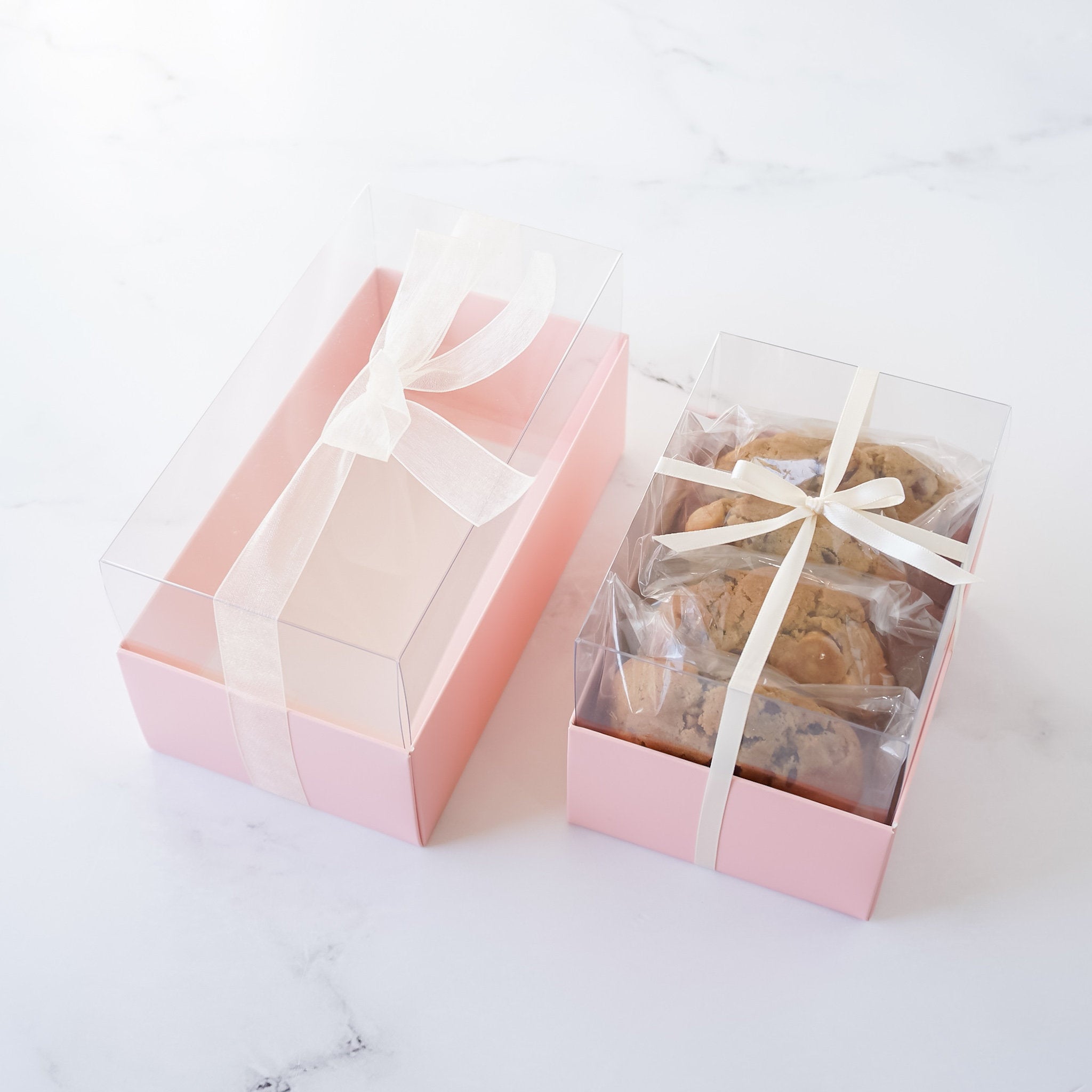 A pink cookie box with a clear lid, containing cookies wrapped in individual clear packaging, tied with a ribbon.