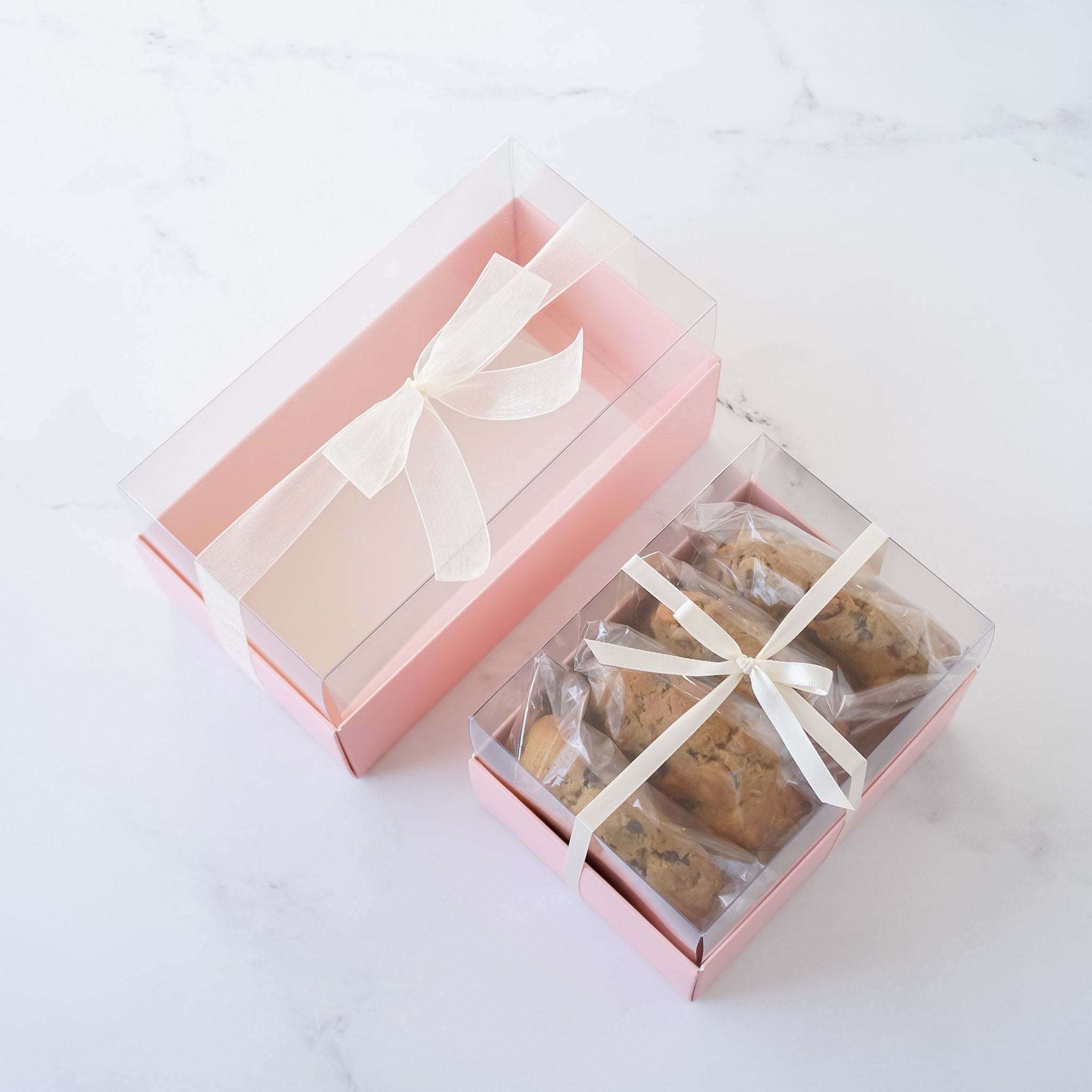 Two pink boxes with clear lids. The boxes are filled with cookies. The cookies are wrapped in clear plastic and tied with white ribbon. The boxes are on a white marble surface.