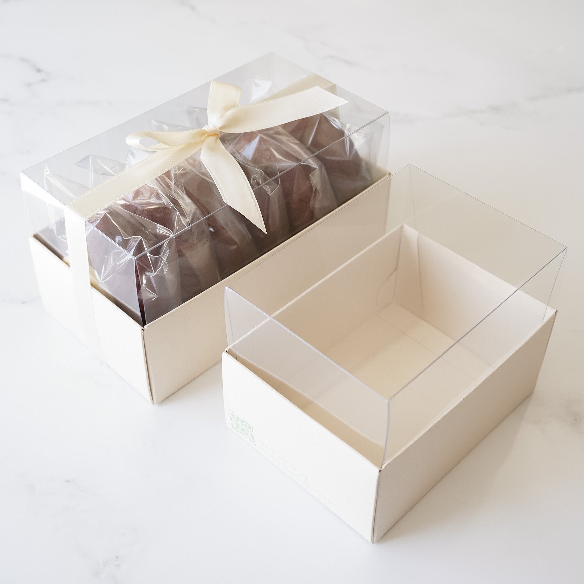 A beige cookie box with a clear lid, containing cookies wrapped in individual clear plastic packaging, tied with a satin bow.